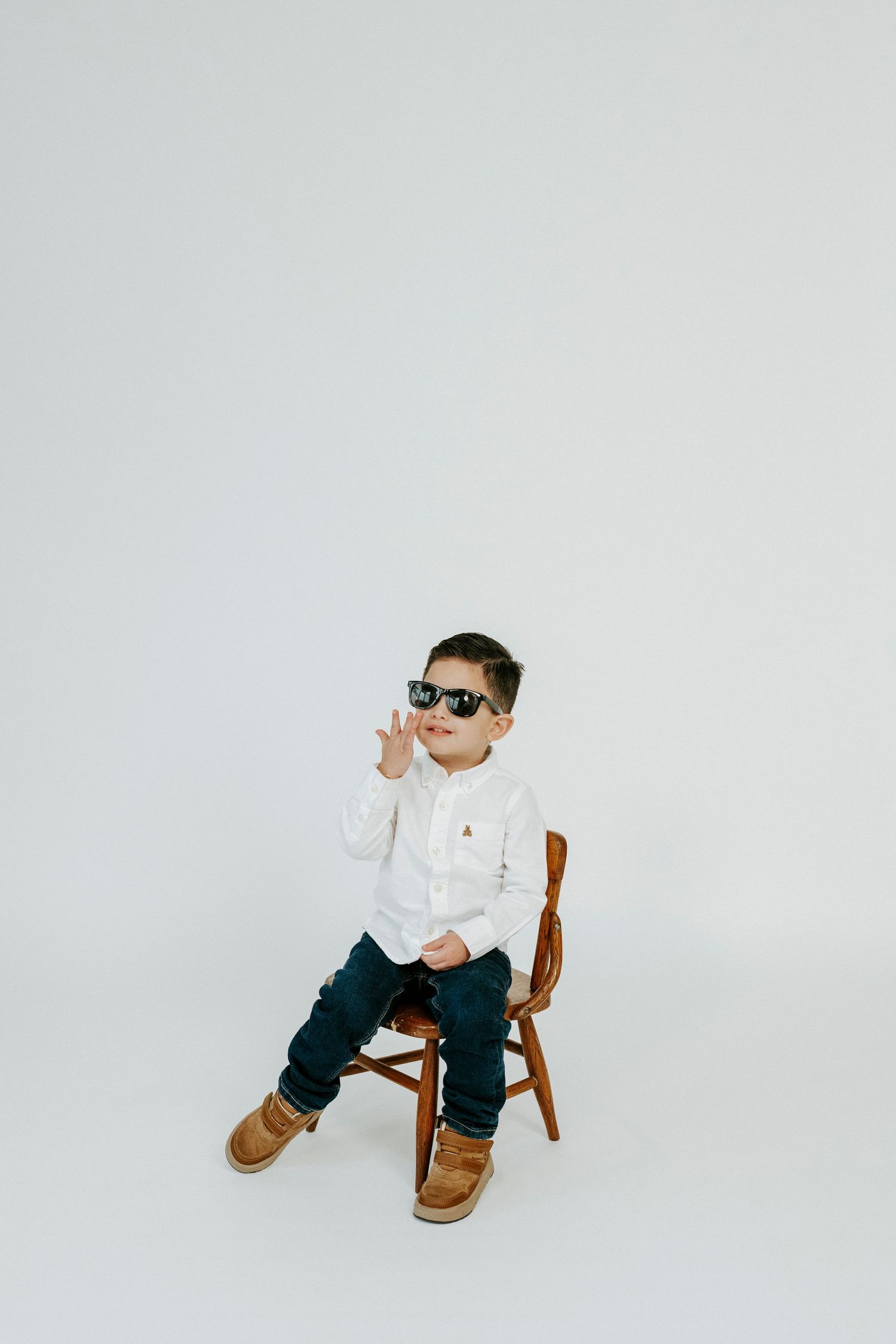 A small child in a white shirt and jeans sits on a wooden chair wearing sunglasses and brown boots against a white backdrop.