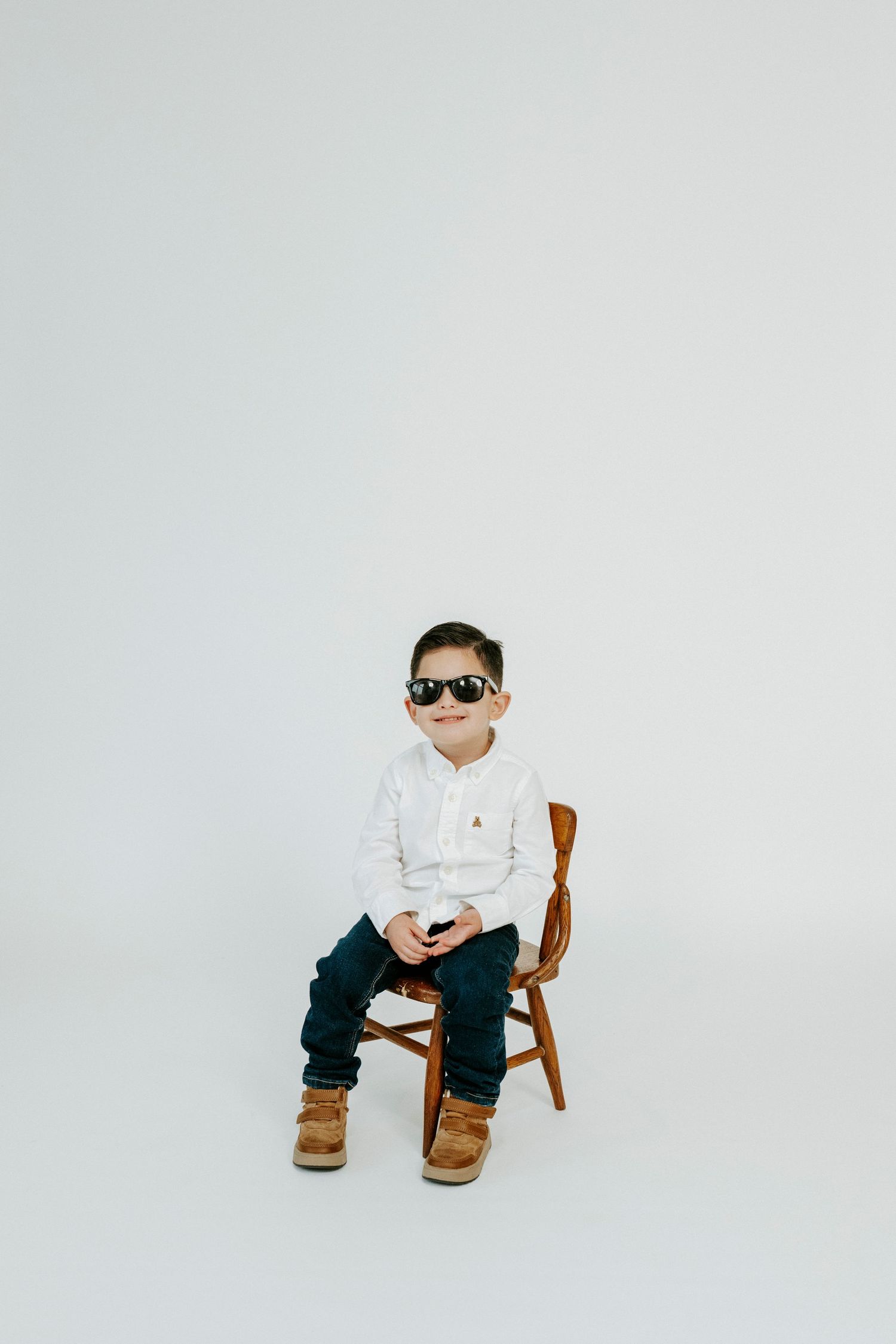 A small child in sunglasses and white shirt sits on a wooden chair against a white backdrop.