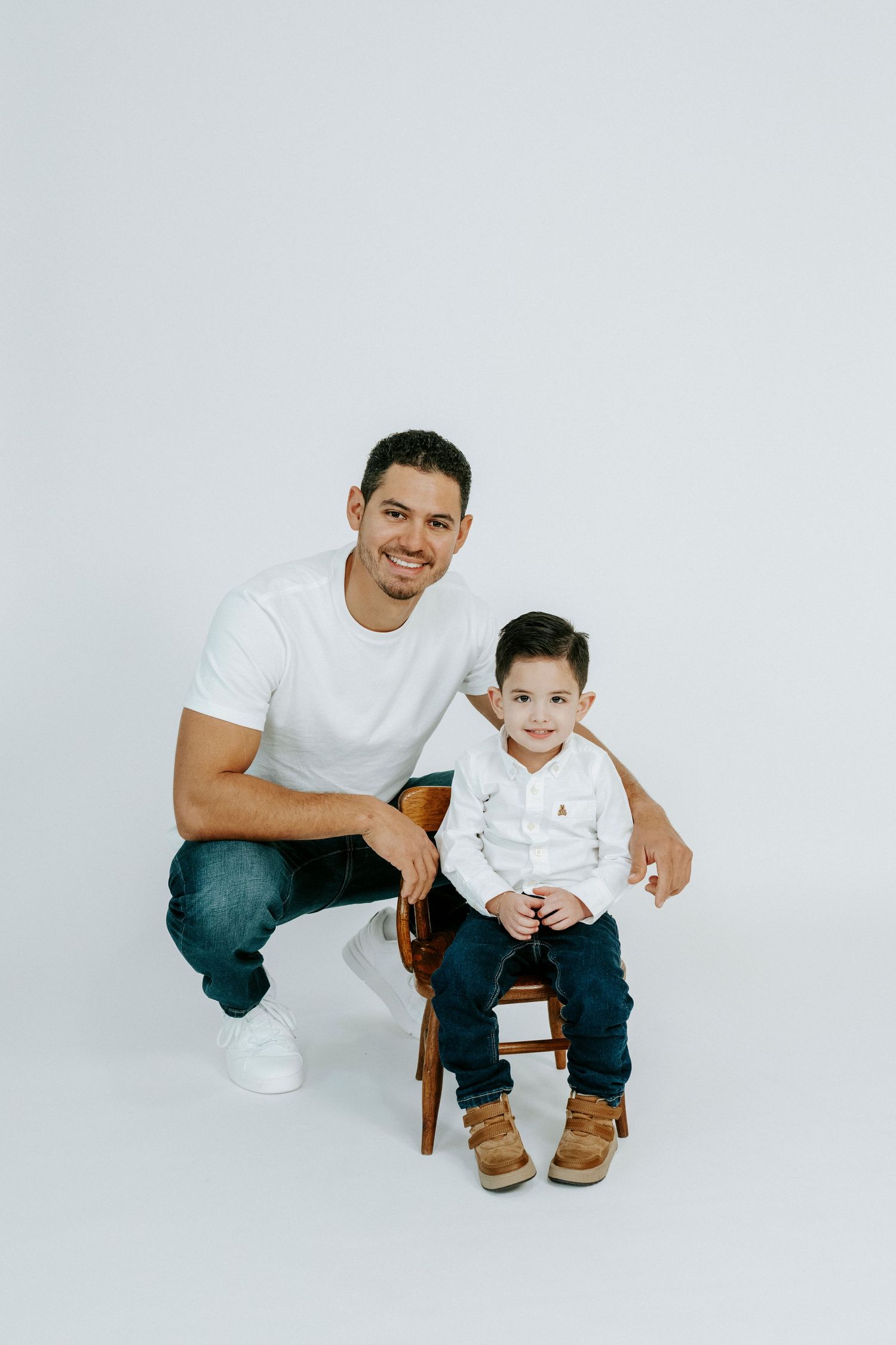 A parent and child sit together on a wooden chair wearing matching white shirts and blue jeans in a minimalist setting.