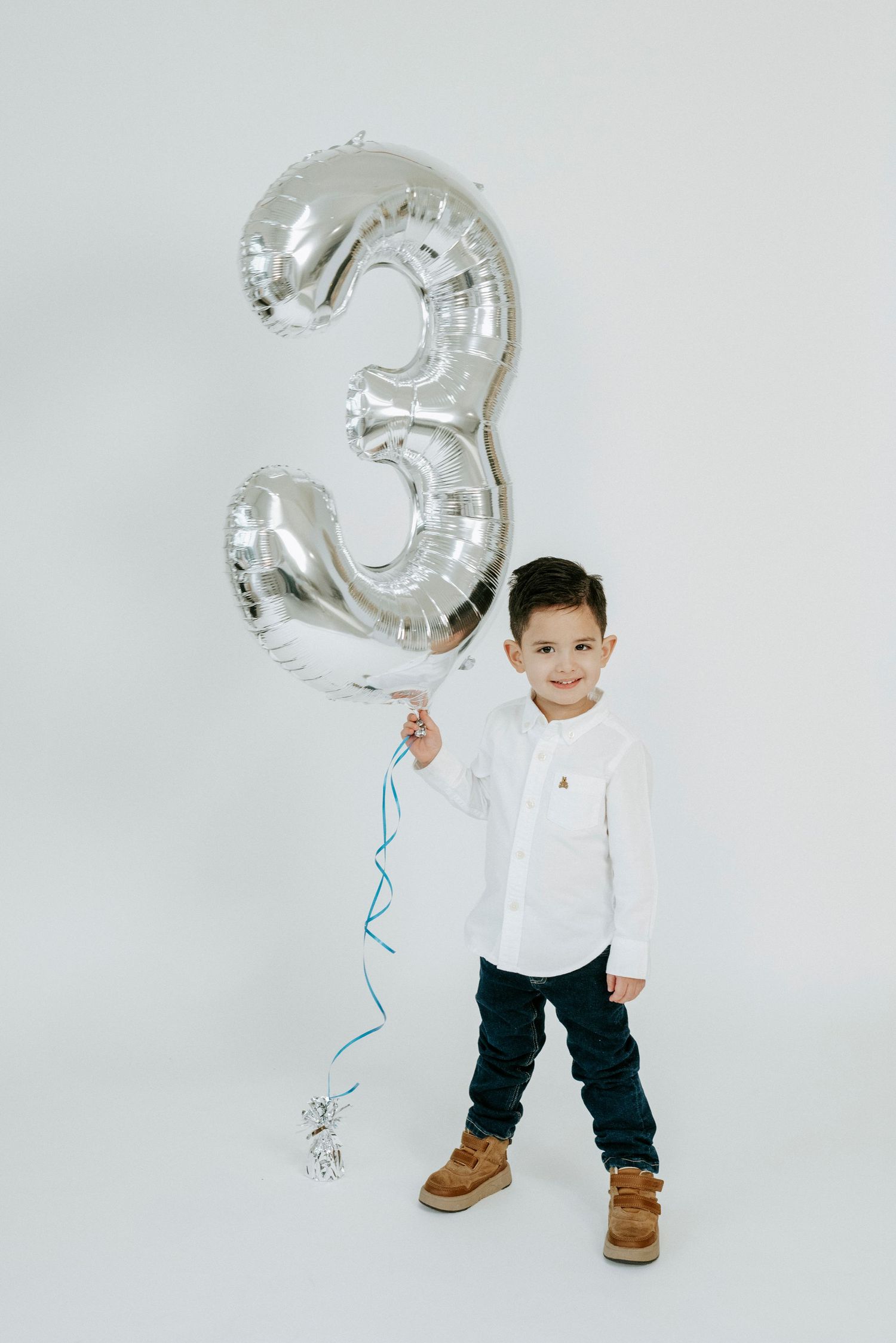A child stands against a white backdrop holding a large silver number 3 balloon while wearing a white shirt and boots.