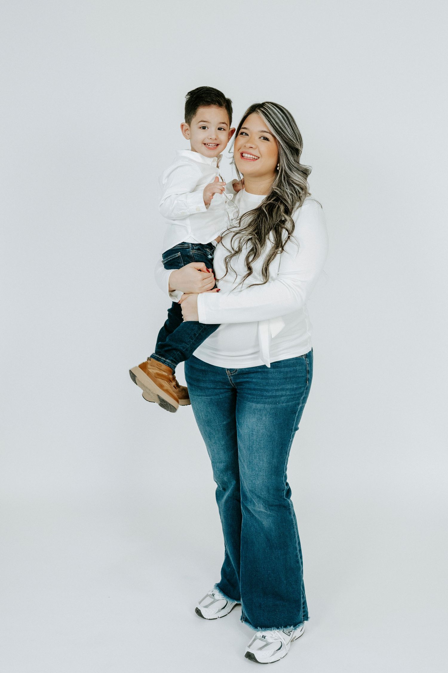 Mother and child in coordinating white and denim outfits pose against white backdrop in lifestyle photoshoot.