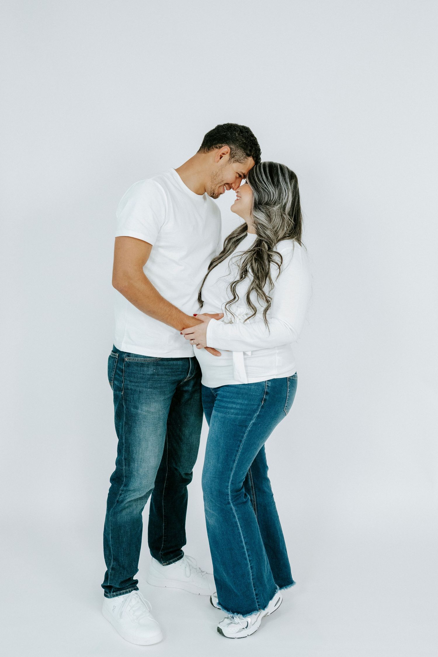 Couple in casual white tops and jeans embrace while laughing against white backdrop.