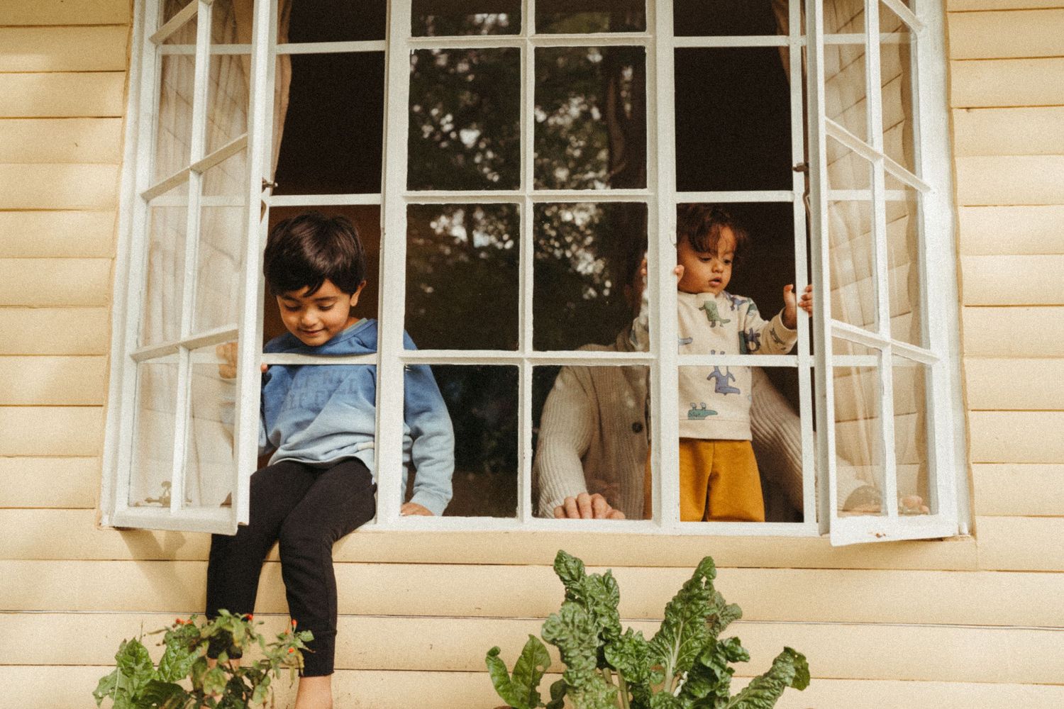Children peek through open windows of a cream-colored cottage on a sunny day, in Johannesburg