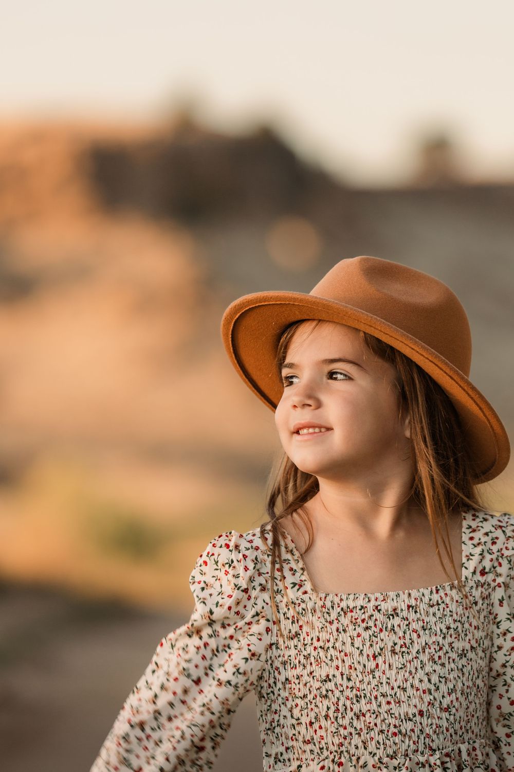 Child in floral dress and brown hat looks up during sunset.