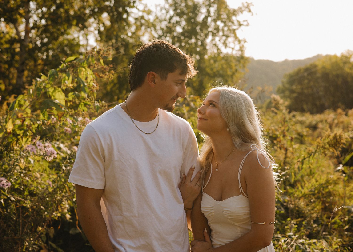 Young couple smiling at each other in a sun-drenched outdoor setting with lush green foliage behind them.