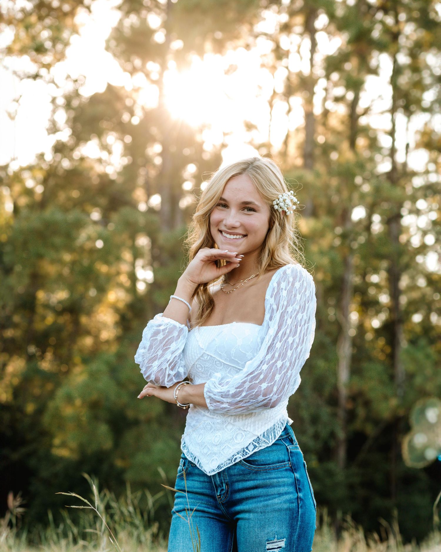 Person in white lace top and blue jeans poses outdoors during golden hour with trees in background.