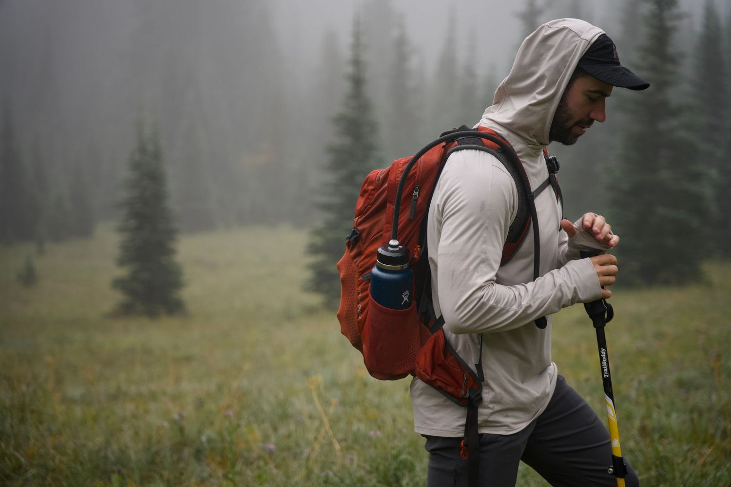 A hiker in a hooded shirt and orange backpack walks through a foggy alpine meadow with trekking poles.