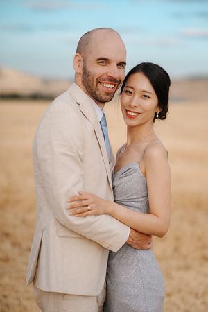 Couple embraces lovingly in a golden field at sunset wearing light colored formal attire.