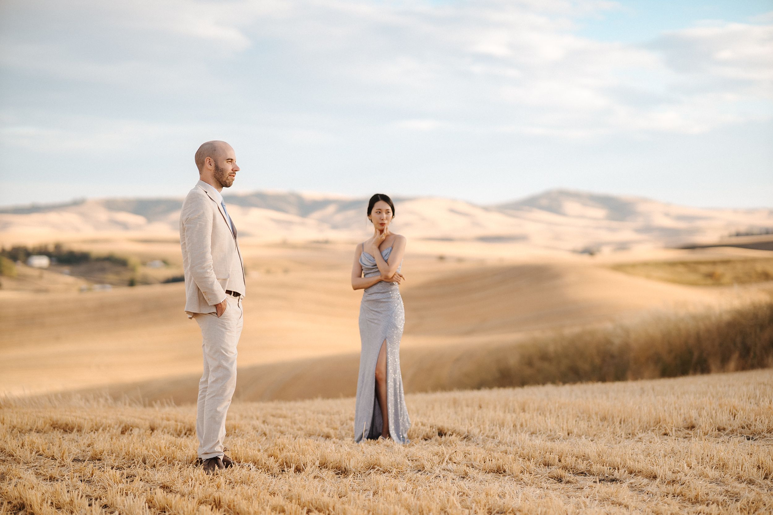 Romantic couple stands together in distance across rolling desert hills during golden hour.