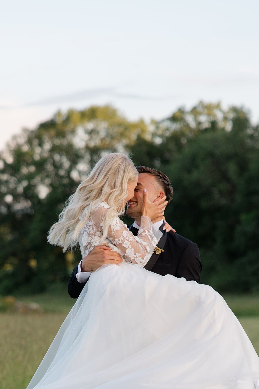 A romantic wedding couple embraces in a sunlit field with trees in the background during golden hour.
