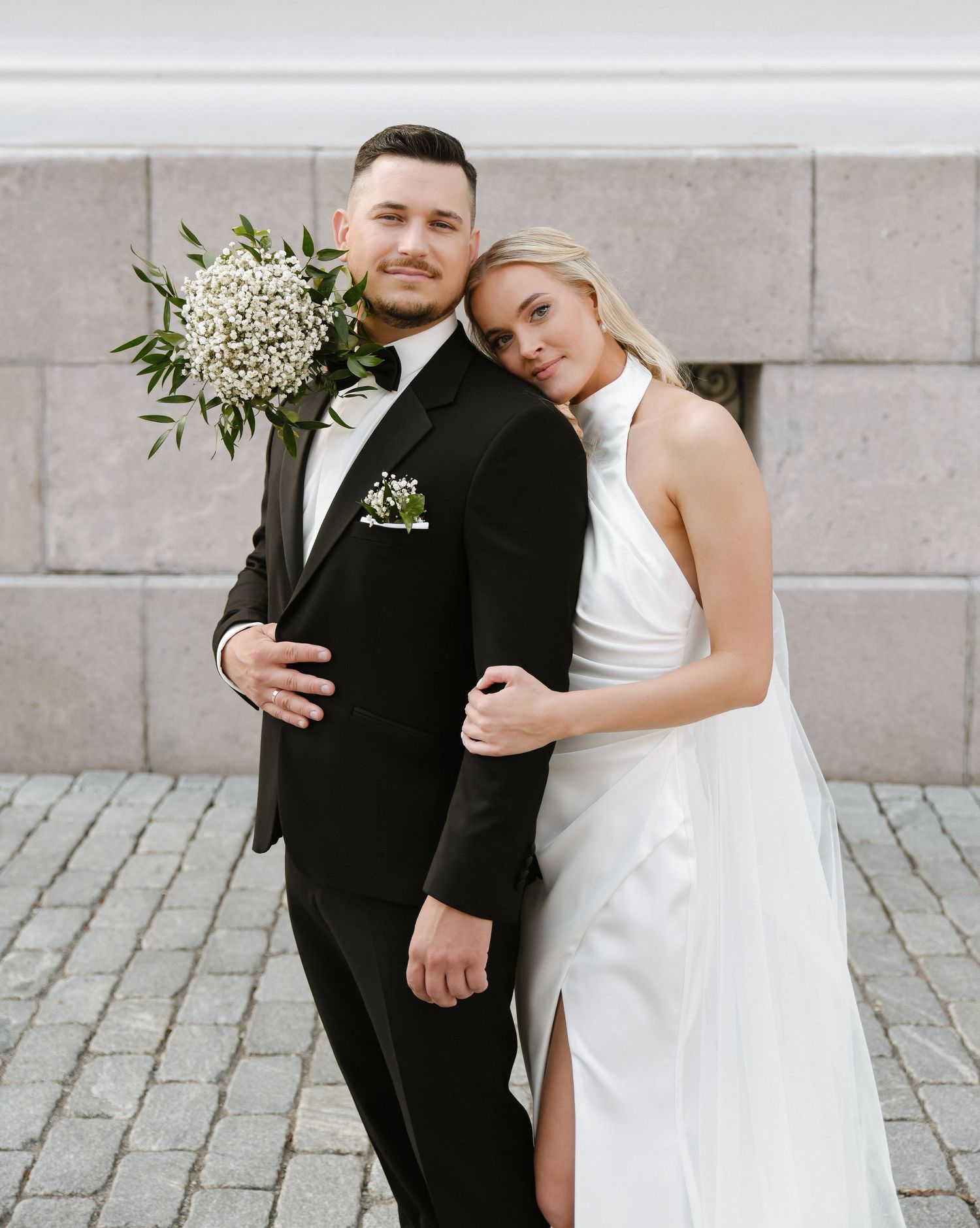A couple in wedding attire embrace outdoors against a stone wall in black and white tones.