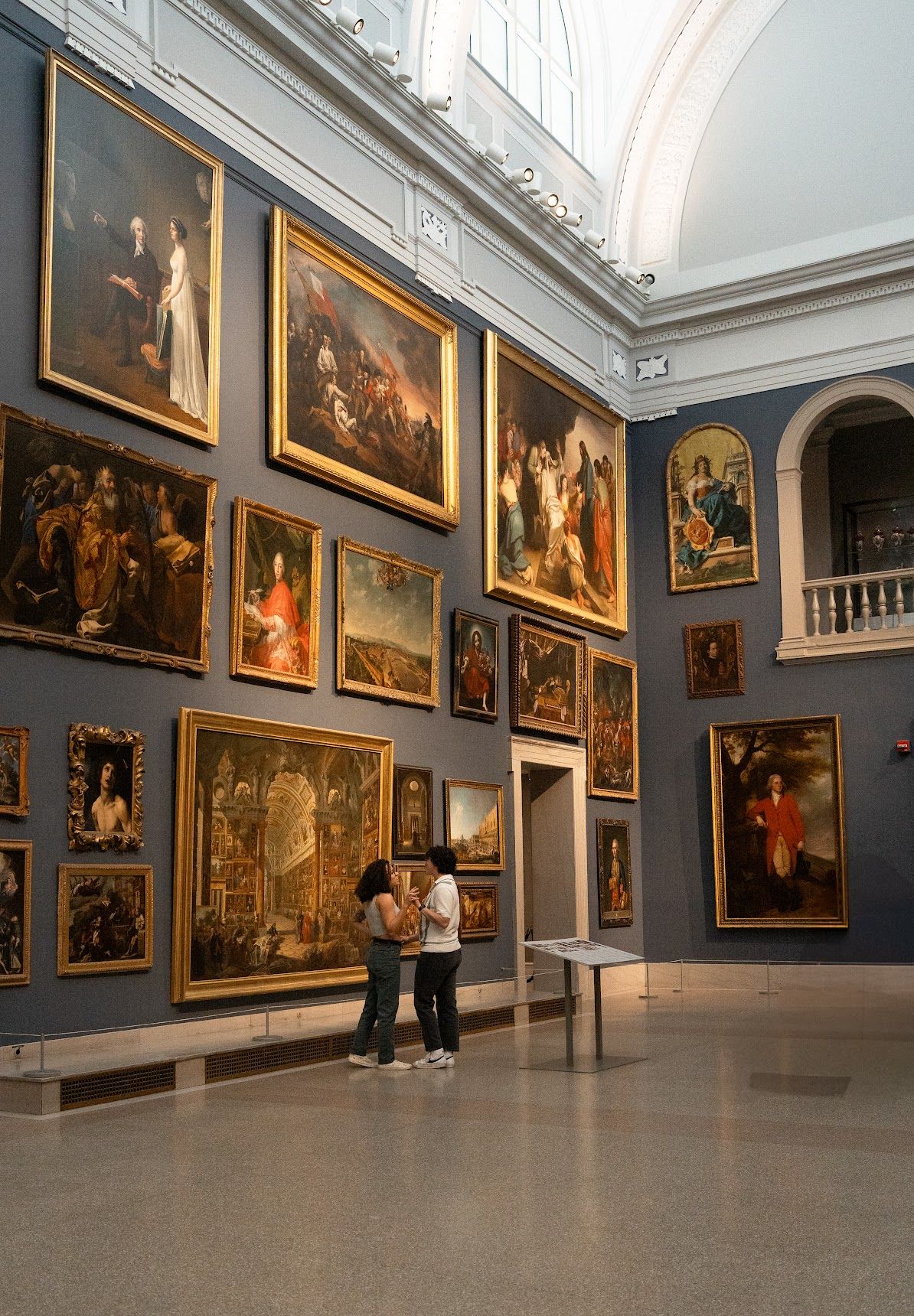 Art gallery interior with classical paintings displayed on gray walls and visitors viewing the exhibition.