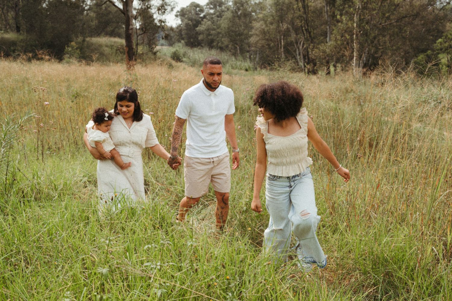 A family wearing white and neutral tones walks together through a grassy field on a sunny day.