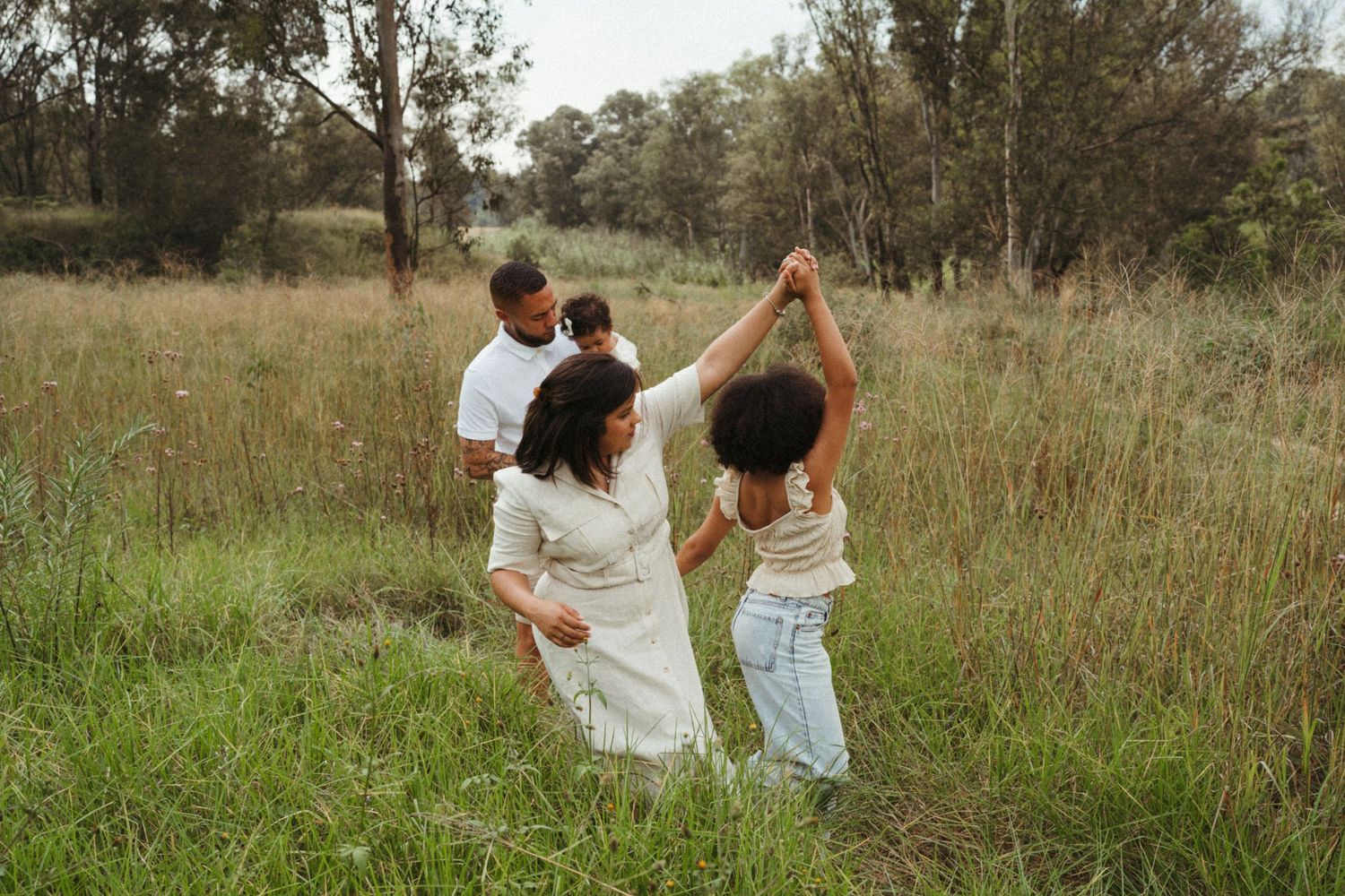 Family members hold hands while walking through tall grass in a natural outdoor setting.