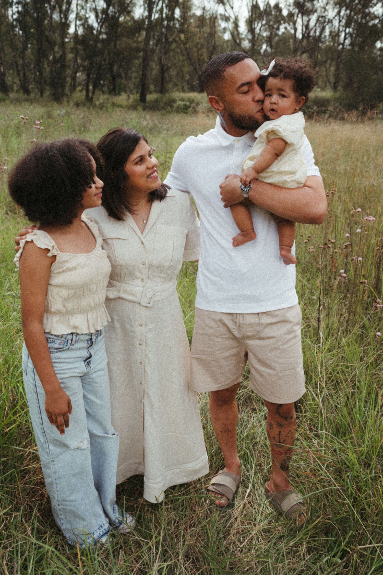 Family dressed in light colors stands together in a field with trees in the background.