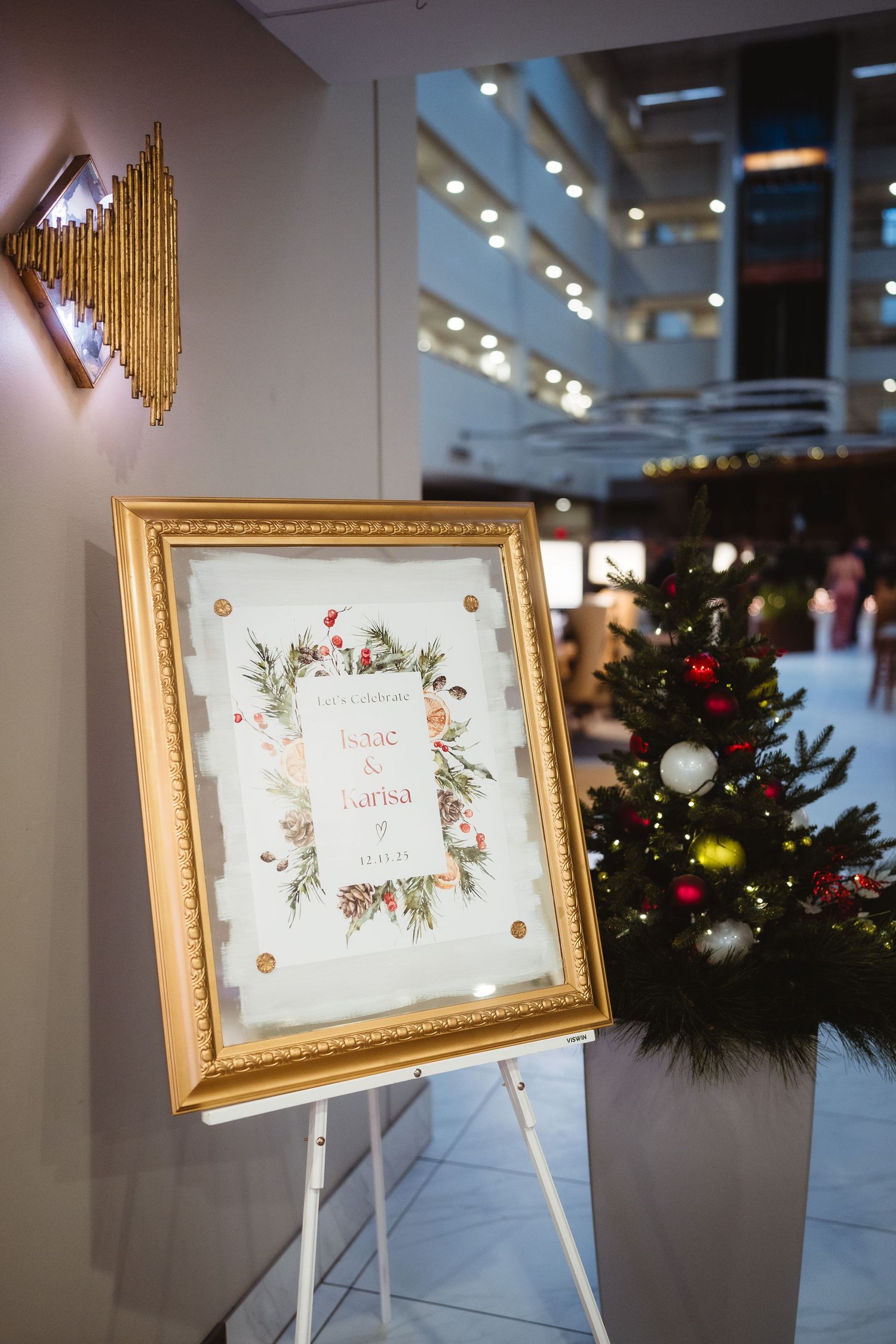 Gold-framed welcome sign with floral design displayed at event venue with Christmas decorations in background.
