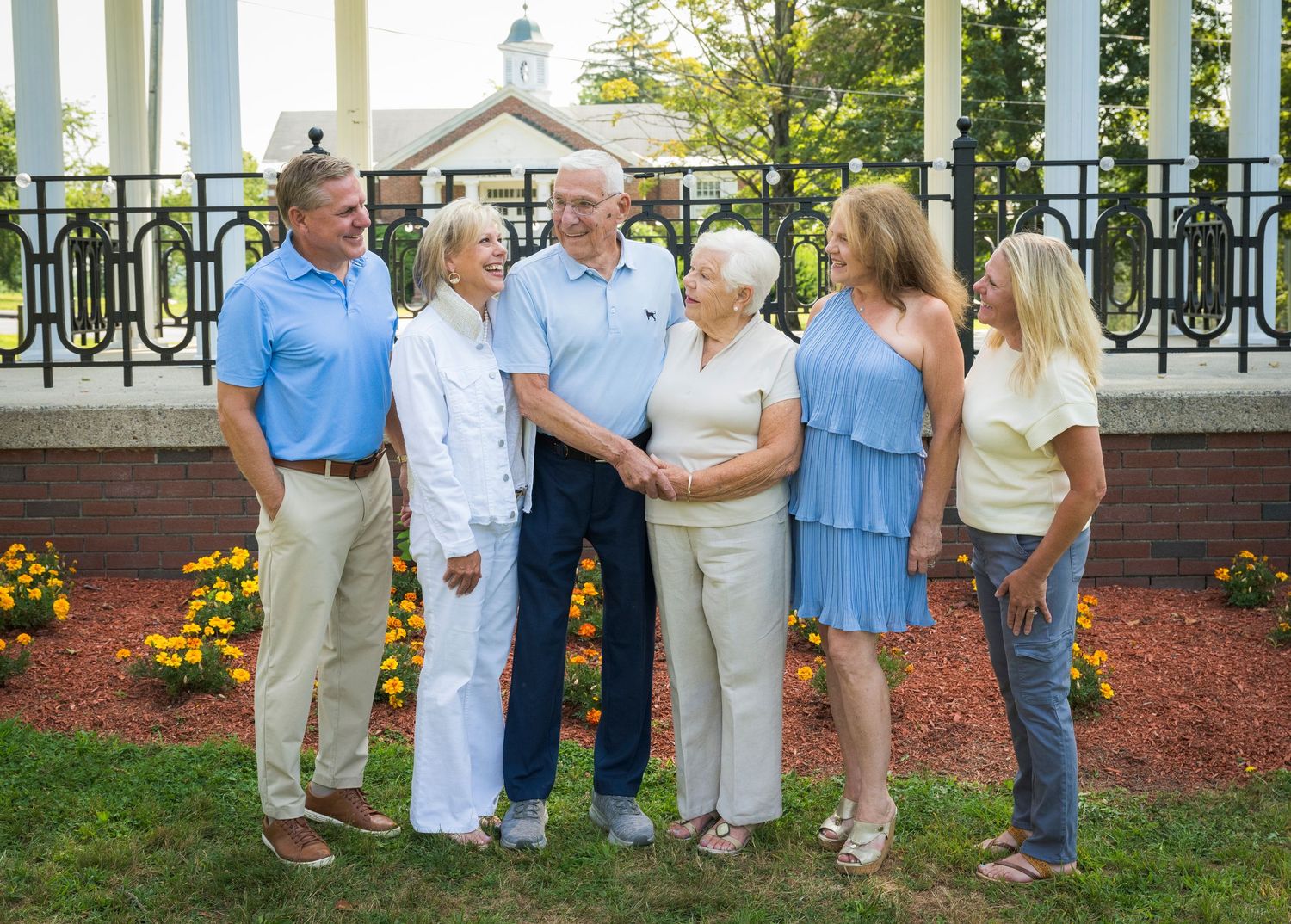 A group of six people stand together outdoors on a summer day, with flowers and ornate railings in the background.