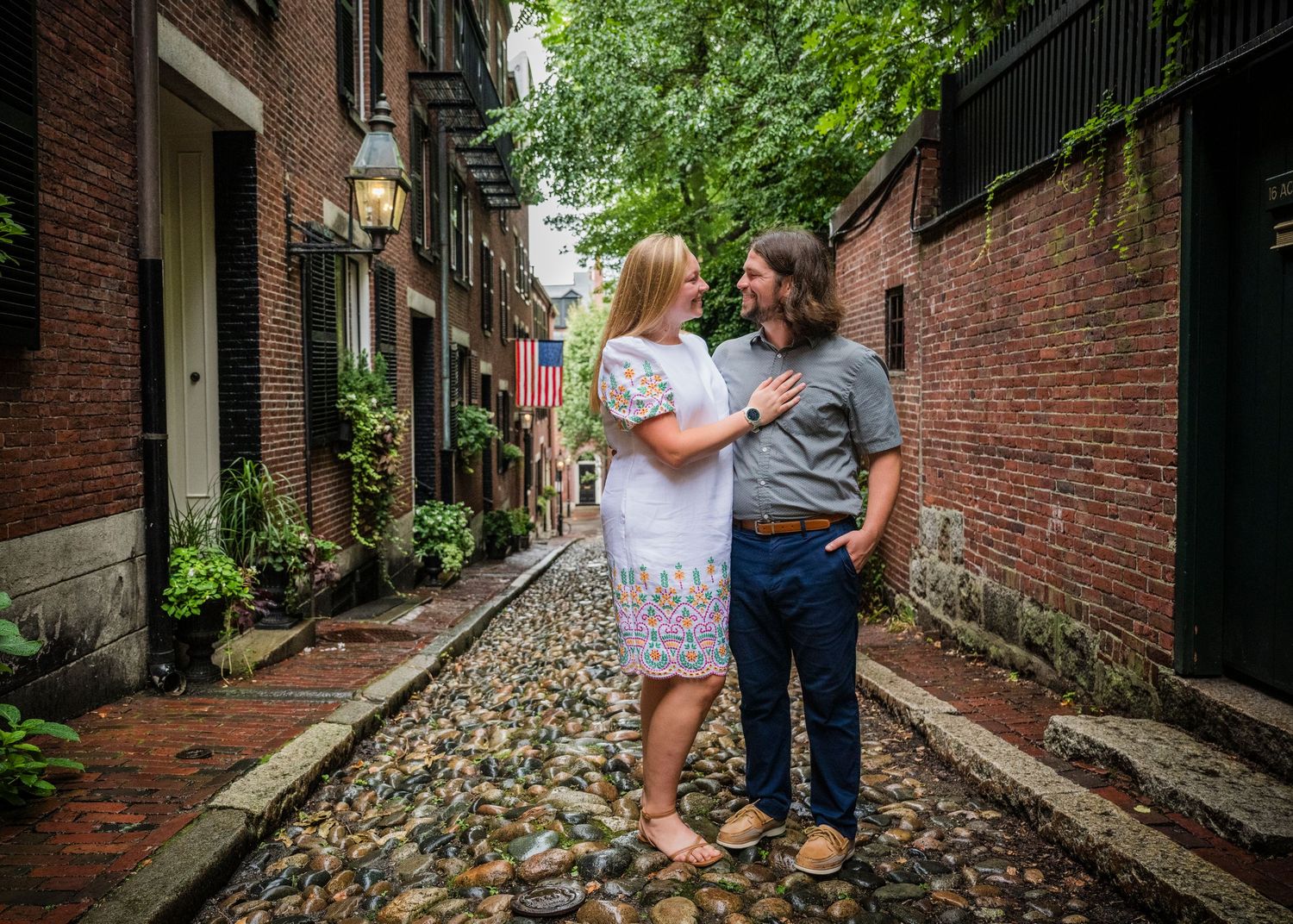 A couple stands together on a charming cobblestone street lined with brick buildings and greenery in Boston's Beacon Hill.