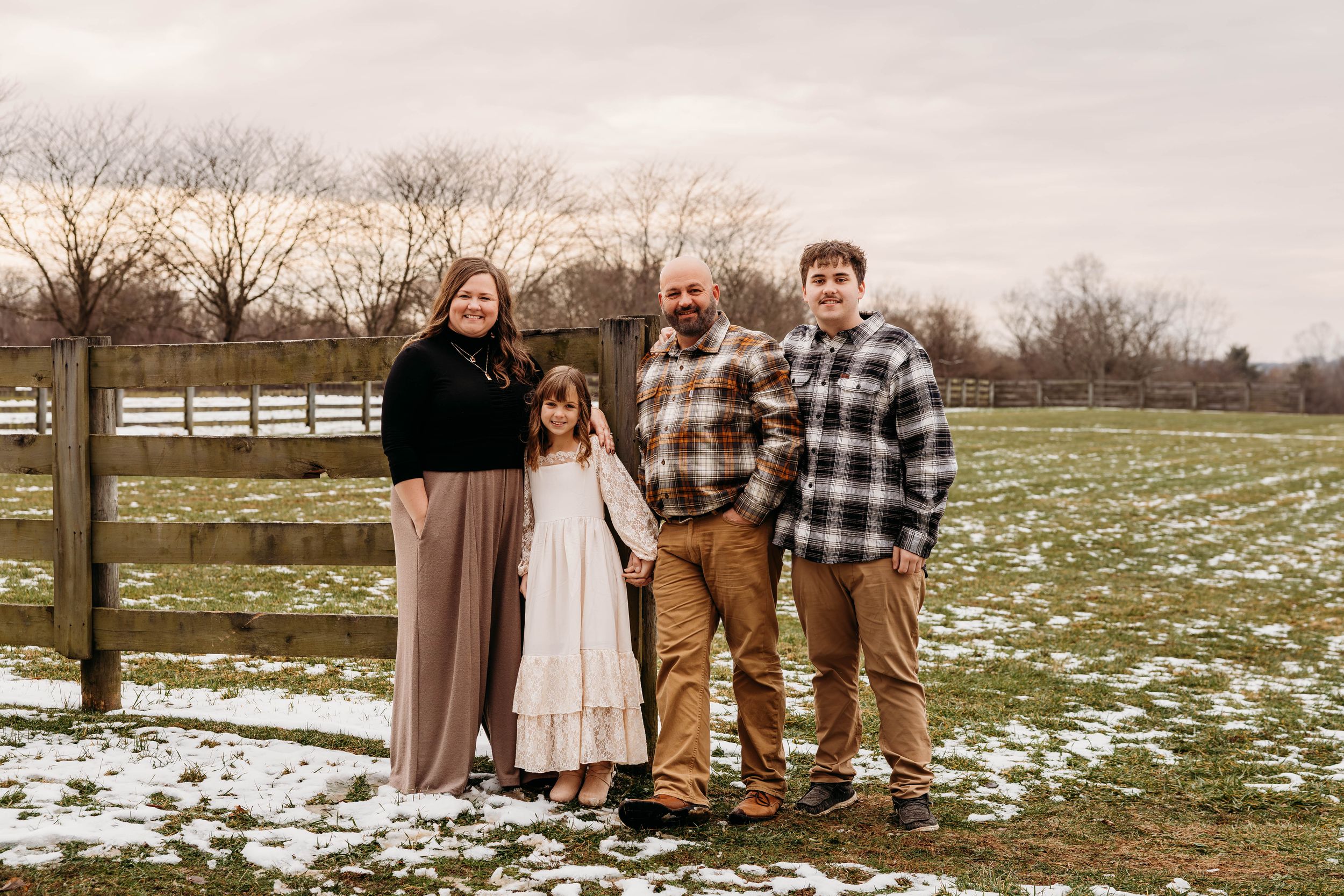 A group stands together in a snowy field with white fencing and bare winter trees in the background.
