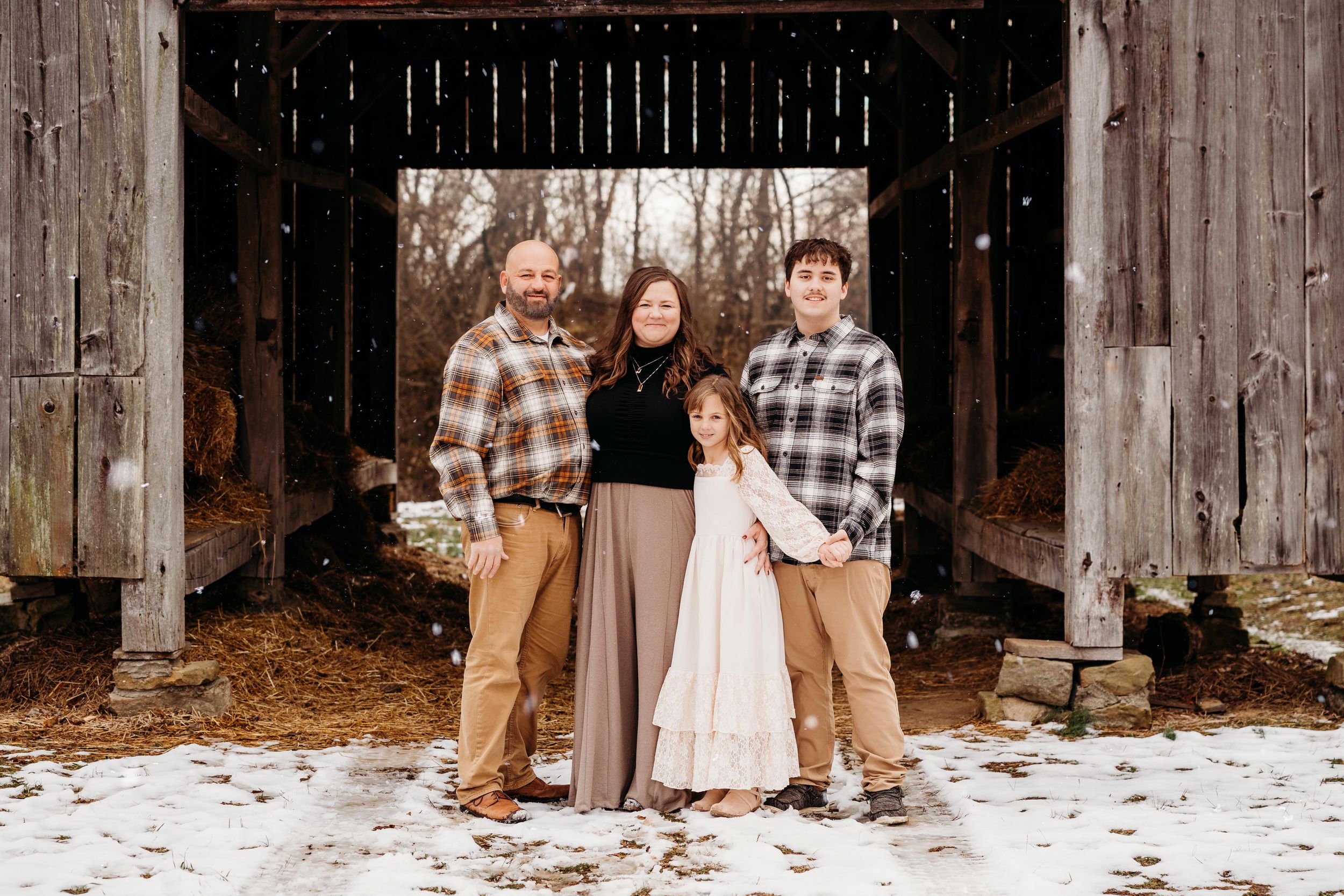 Family in winter outfits poses together in front of rustic wooden barn wall during snowy outdoor photo session.