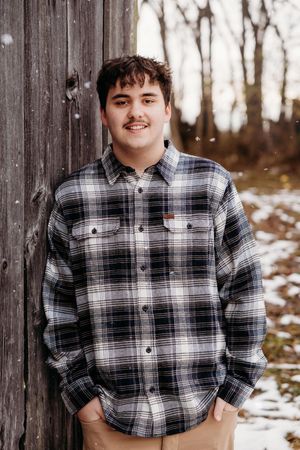 Person in a plaid flannel shirt poses against a wooden fence with snow-covered ground and winter trees in background.