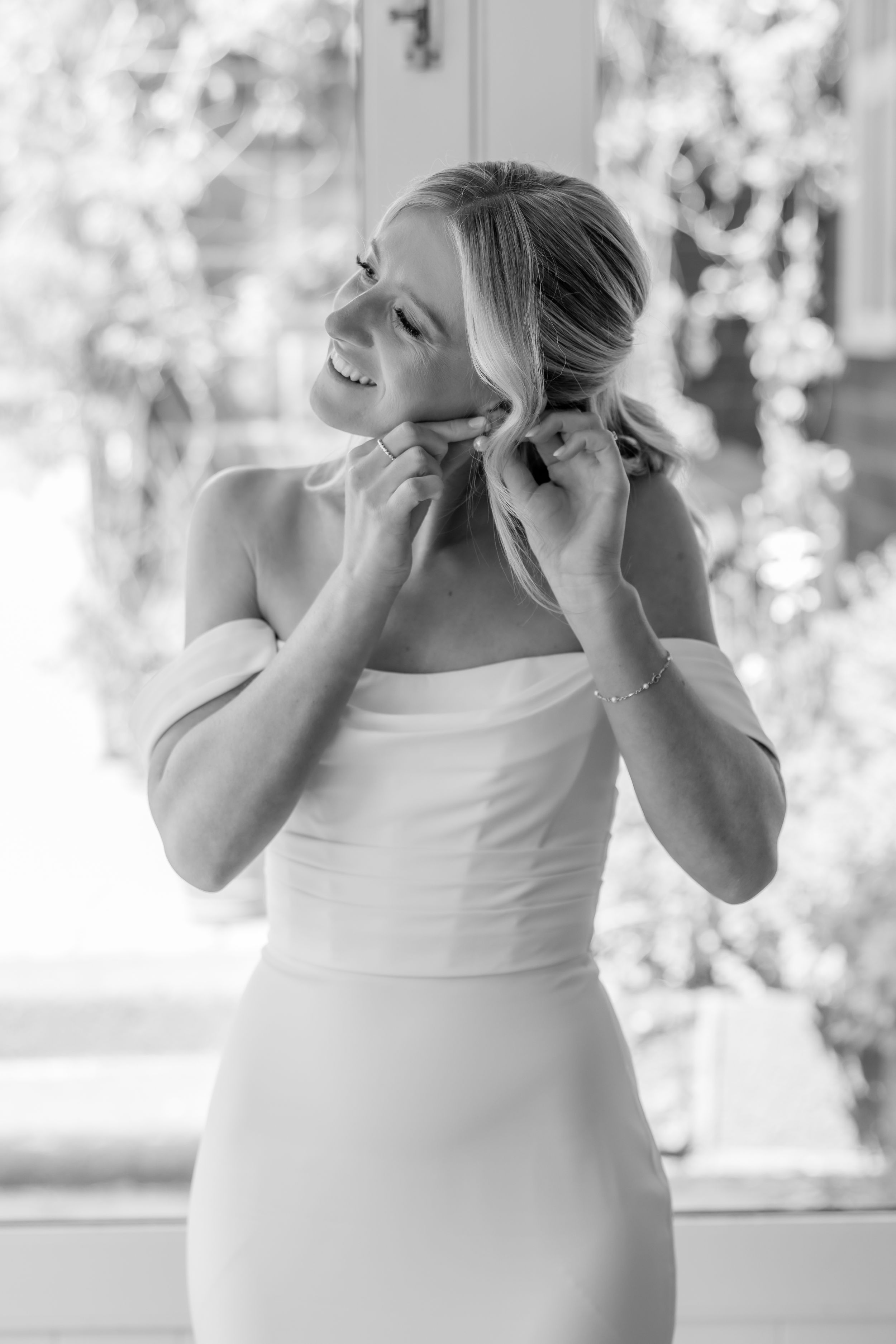 A black and white portrait of someone putting on earrings while wearing a strapless dress.