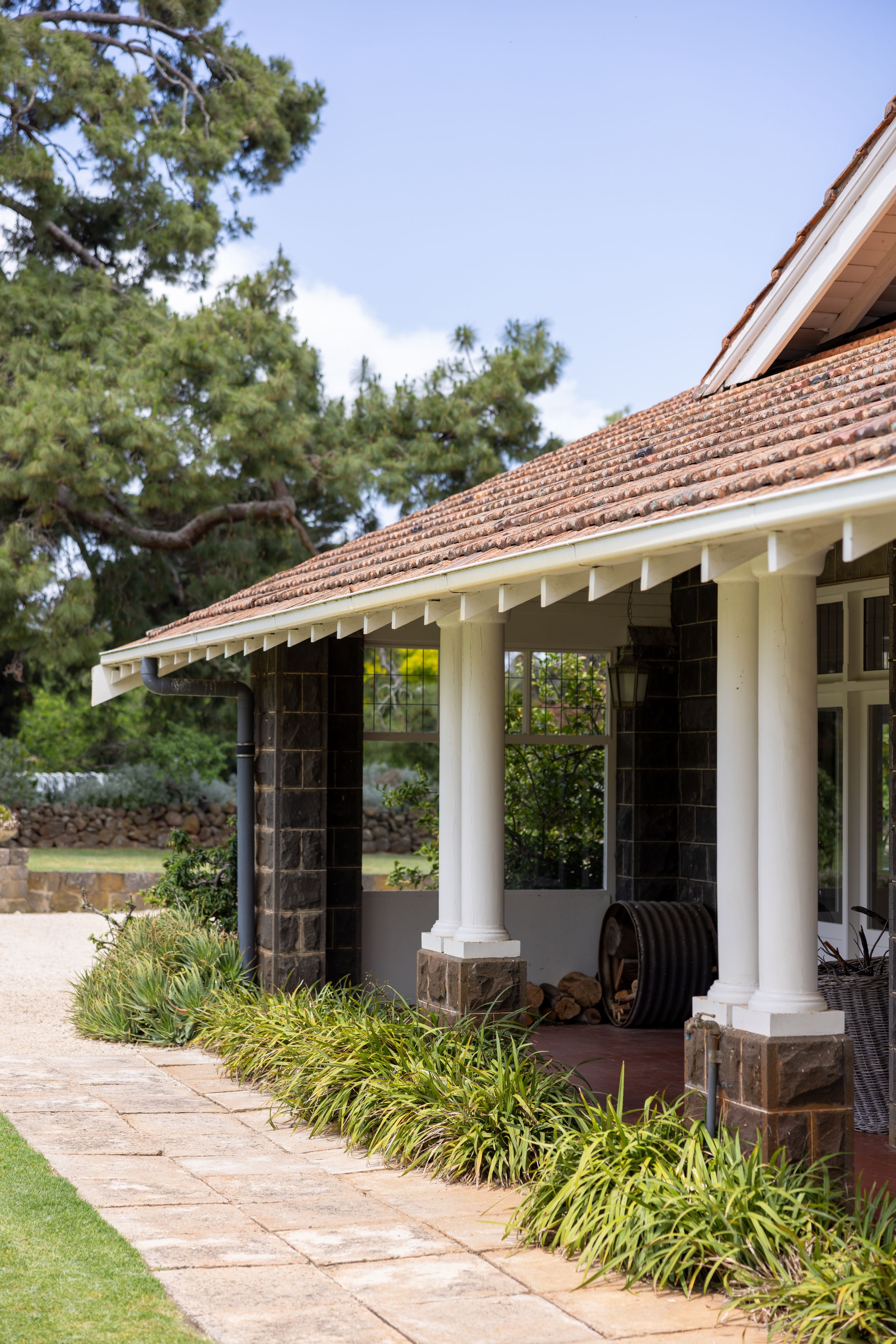 White colonial style porch with stone pillars and terracotta roof tiles overlooking a manicured garden path.
