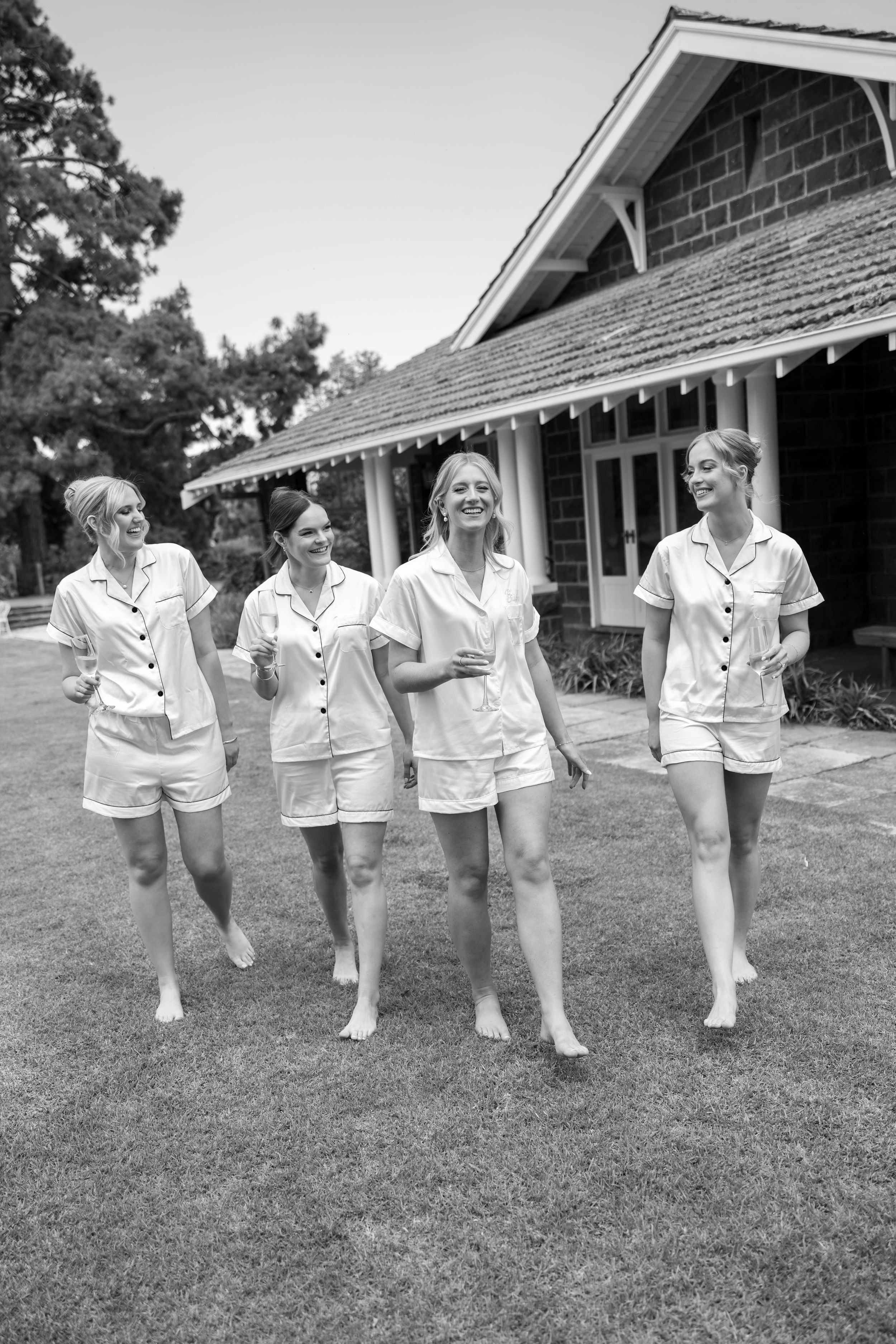 Black and white photo of bridesmaids in matching white outfits dancing in a line on grass.
