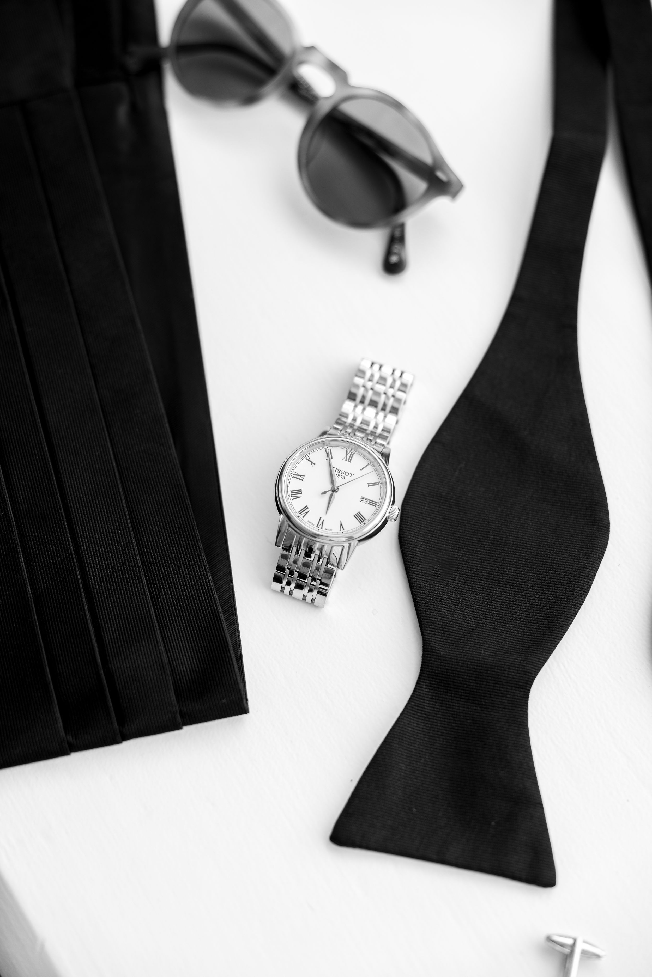 Black and white still life of groom's accessories including bow tie, watch, and sunglasses.