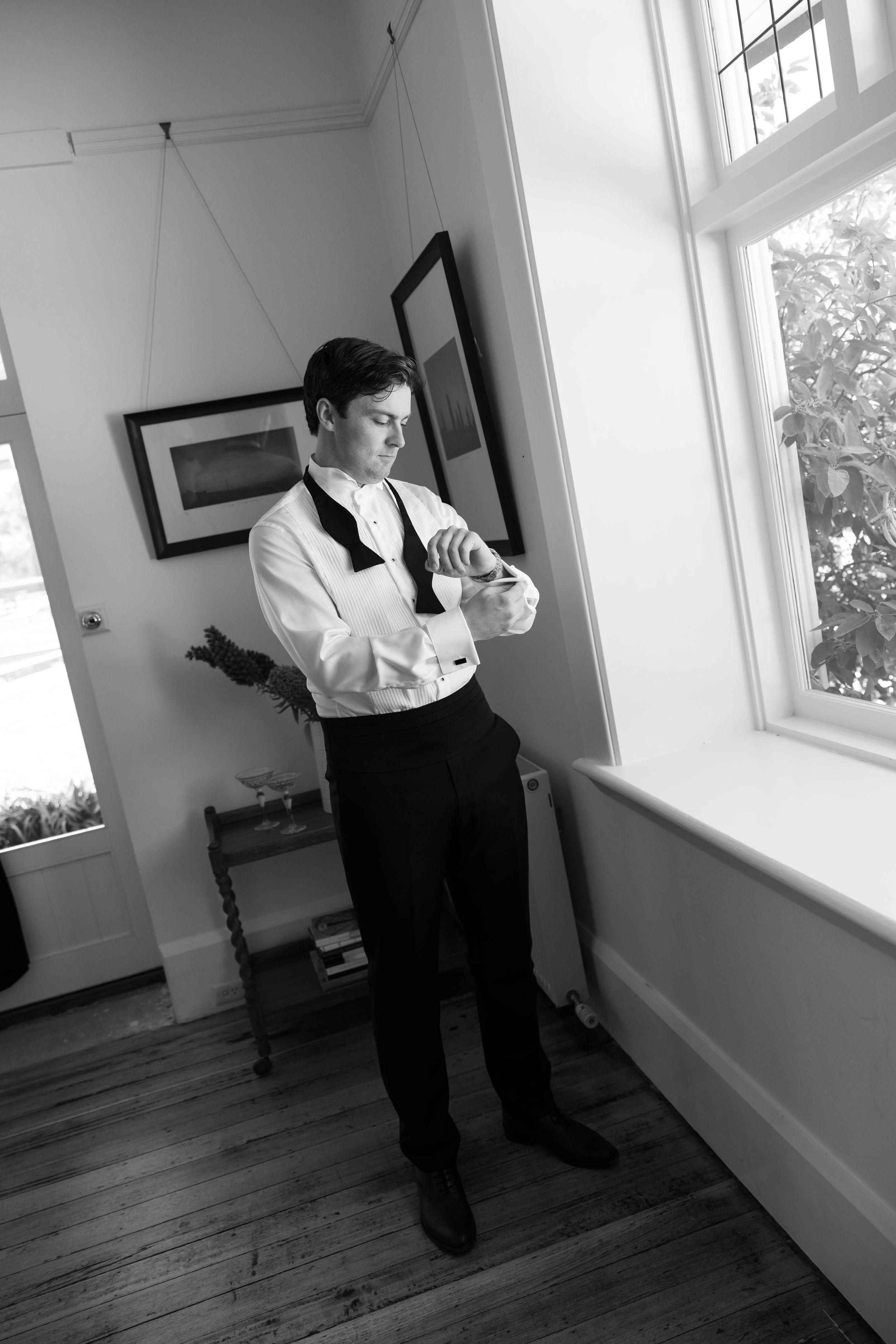 A black and white photo of someone adjusting their tie while getting ready in a sunlit room.