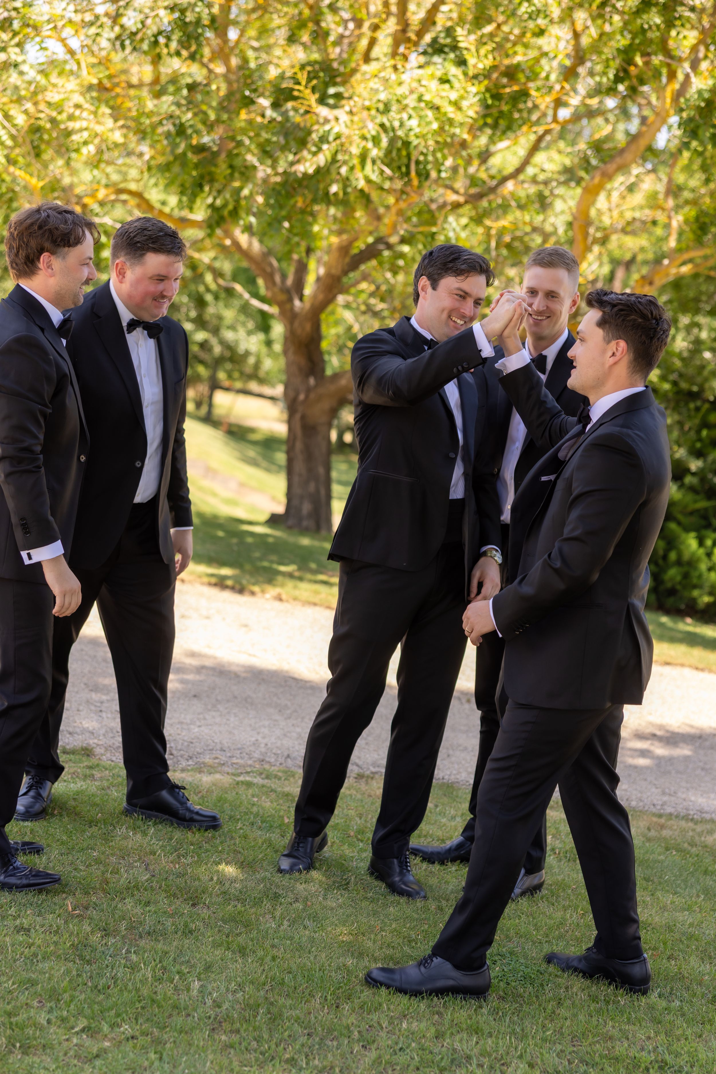 Groomsmen in black tuxedos sharing a laugh on grassy lawn.