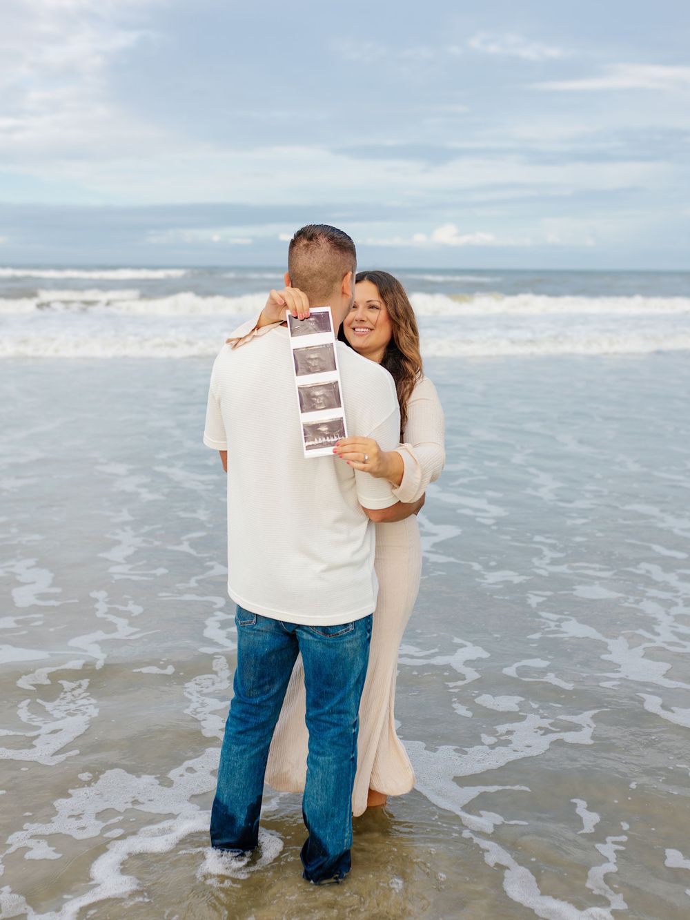 Couple embracing on beach while holding ultrasound photos with ocean waves in background.
