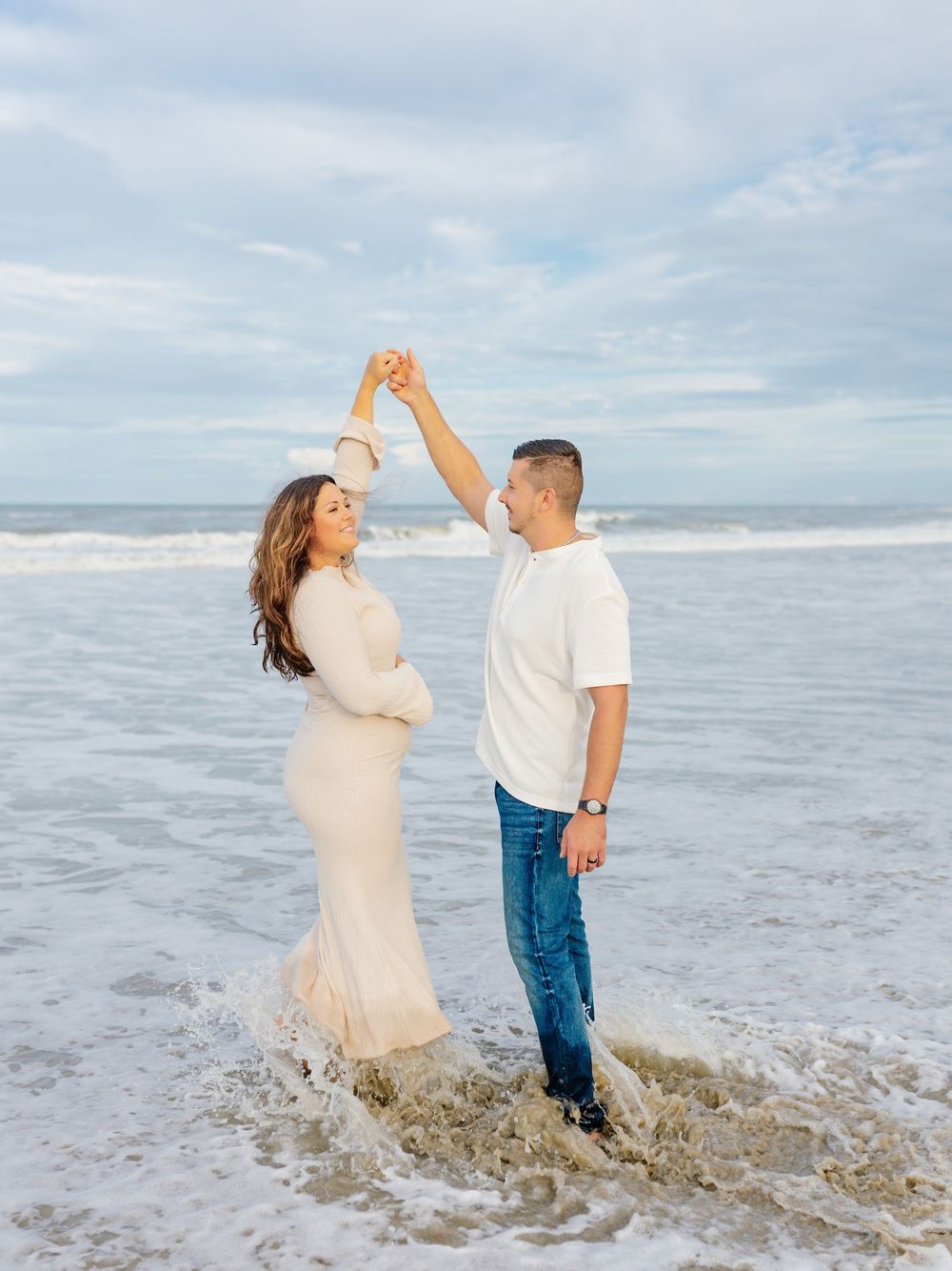 Couple dancing joyfully in ocean waves at sunset during beach photoshoot.
