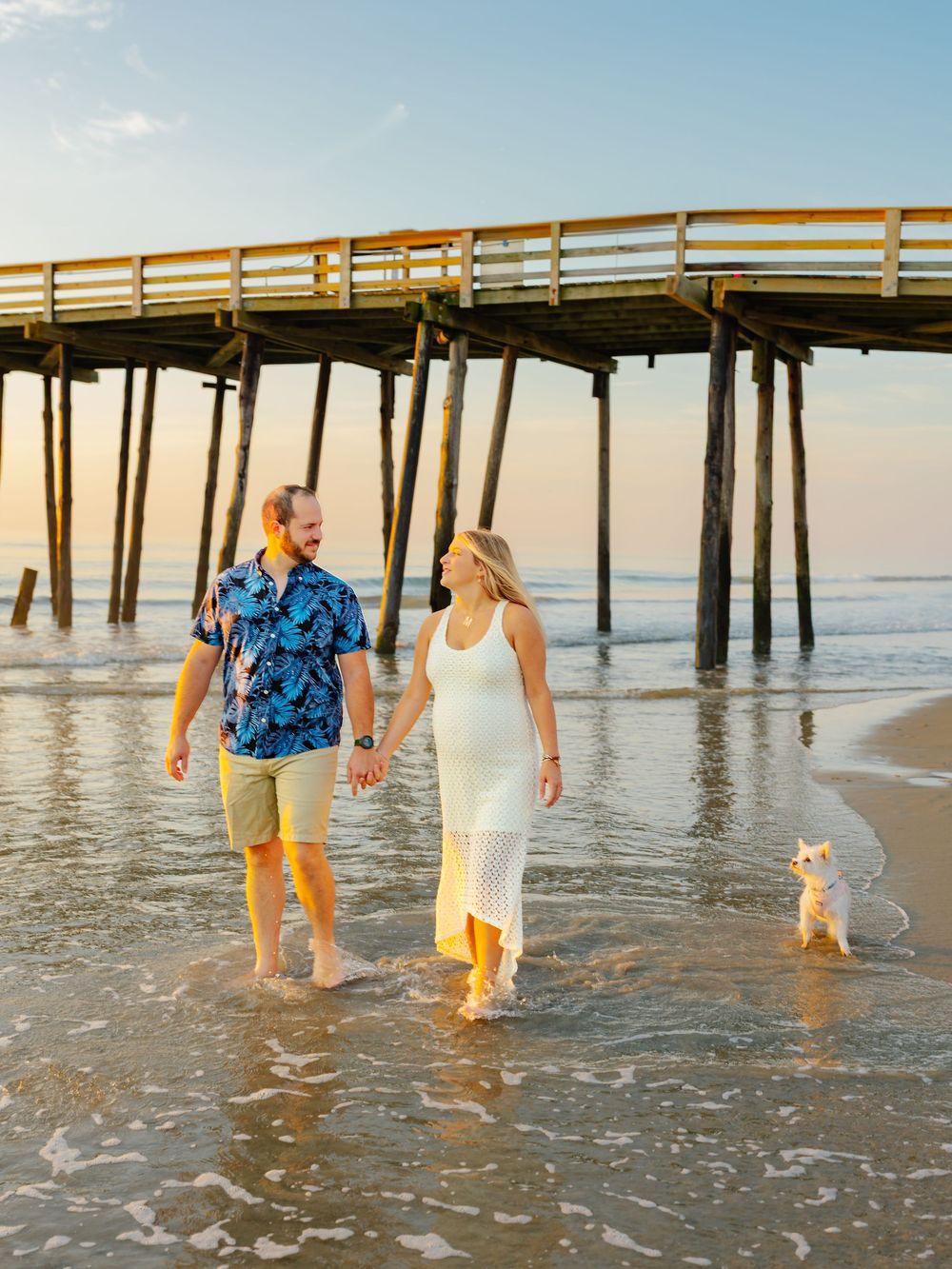 Couple walking through shallow ocean water near wooden pier with dog.