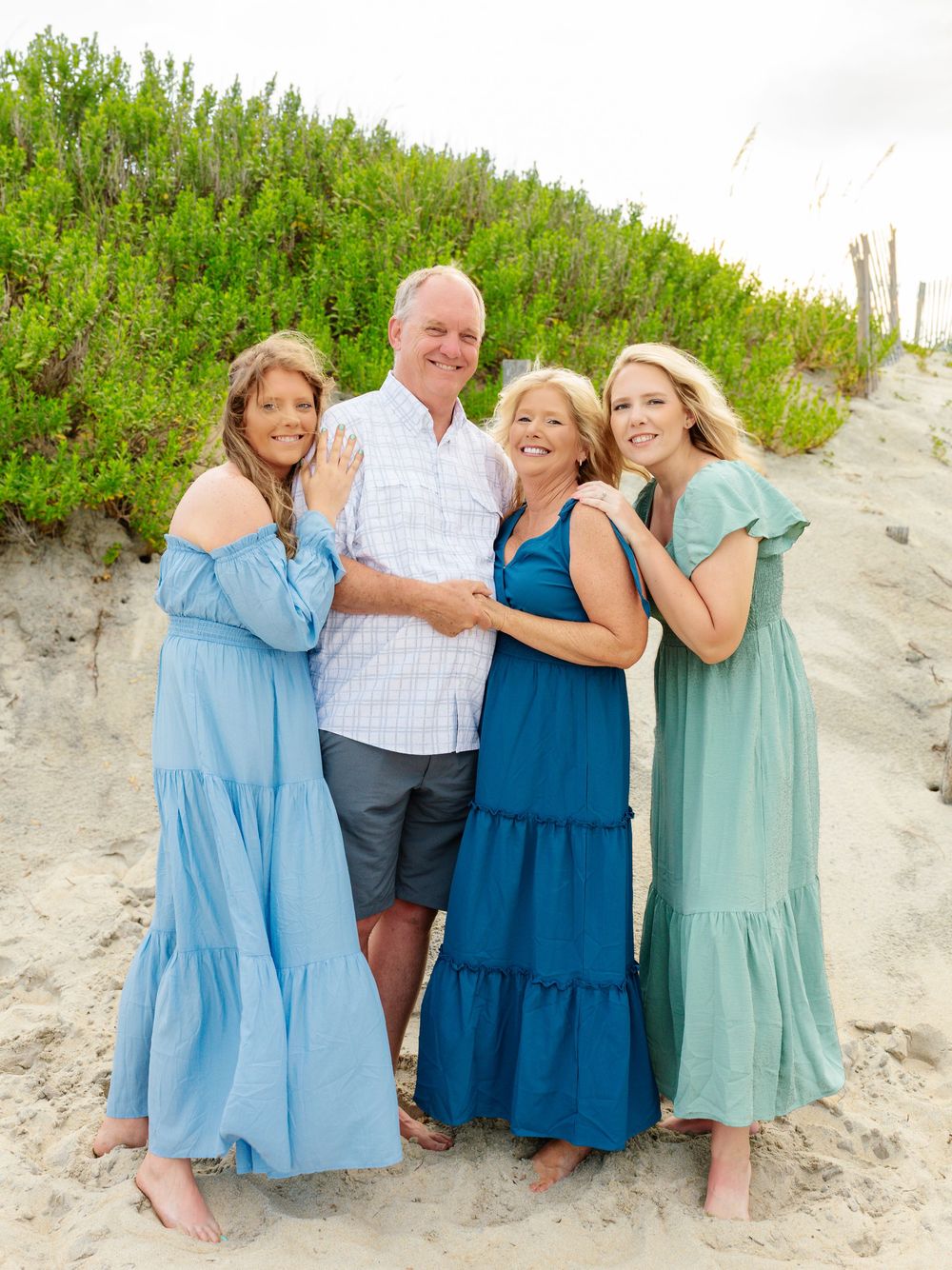Family members in coordinating blue and teal dresses pose on beach dunes.