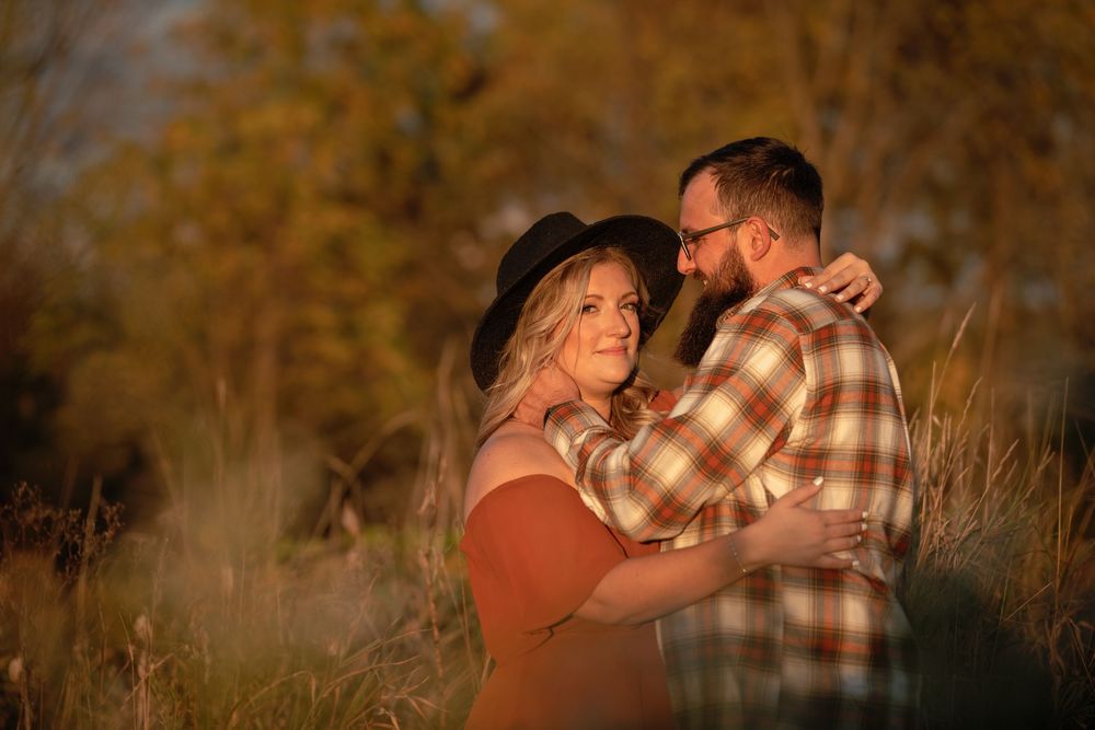 A couple embraces during a romantic sunset photoshoot in a rustic outdoor setting with golden light filtering through trees.