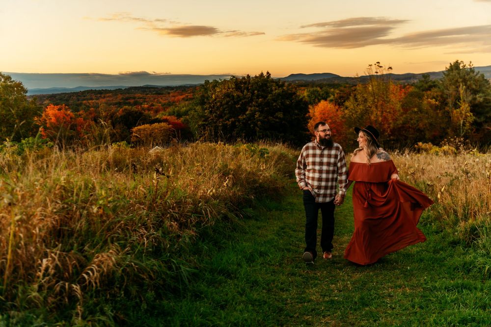 Two figures stand on a green path at sunset with one wearing a flowing red dress against an autumn landscape.