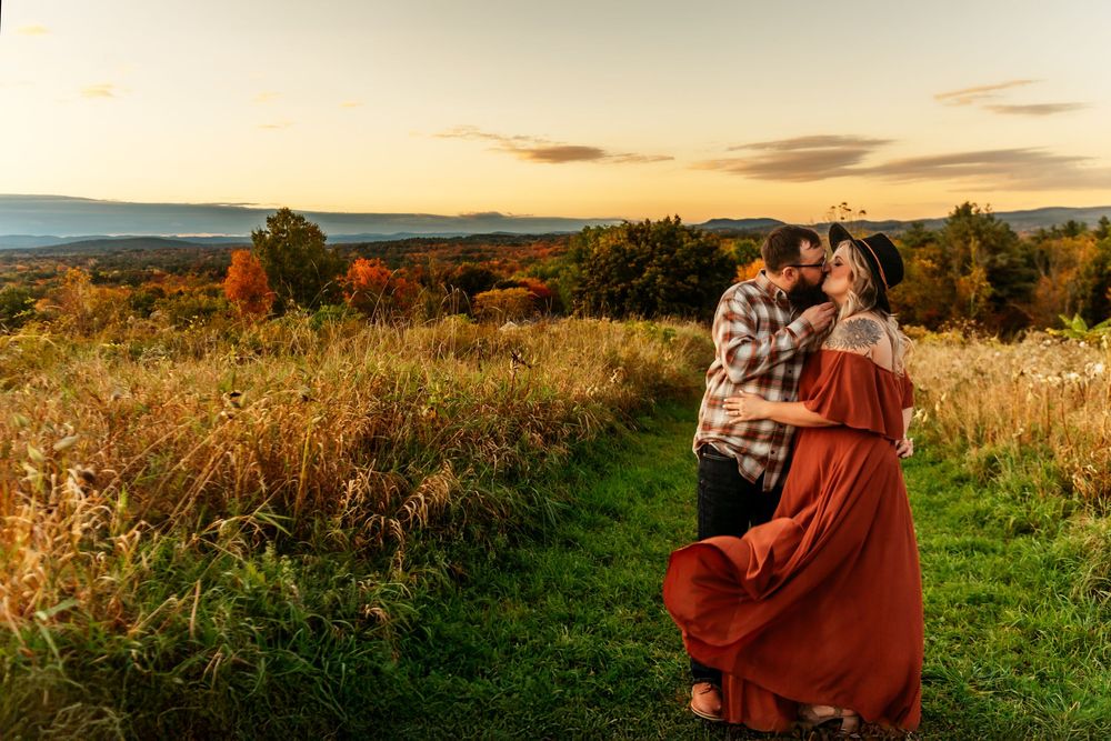 A couple embraces on a grassy hilltop path during golden hour with autumn foliage and mountains in the distance.