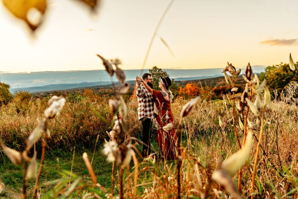 A couple and their dogs enjoy a sunset walk through a rustic field with mountains in the distance.