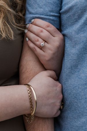 Close-up of hands embracing showing an engagement ring and gold bracelet against blue and brown clothing.