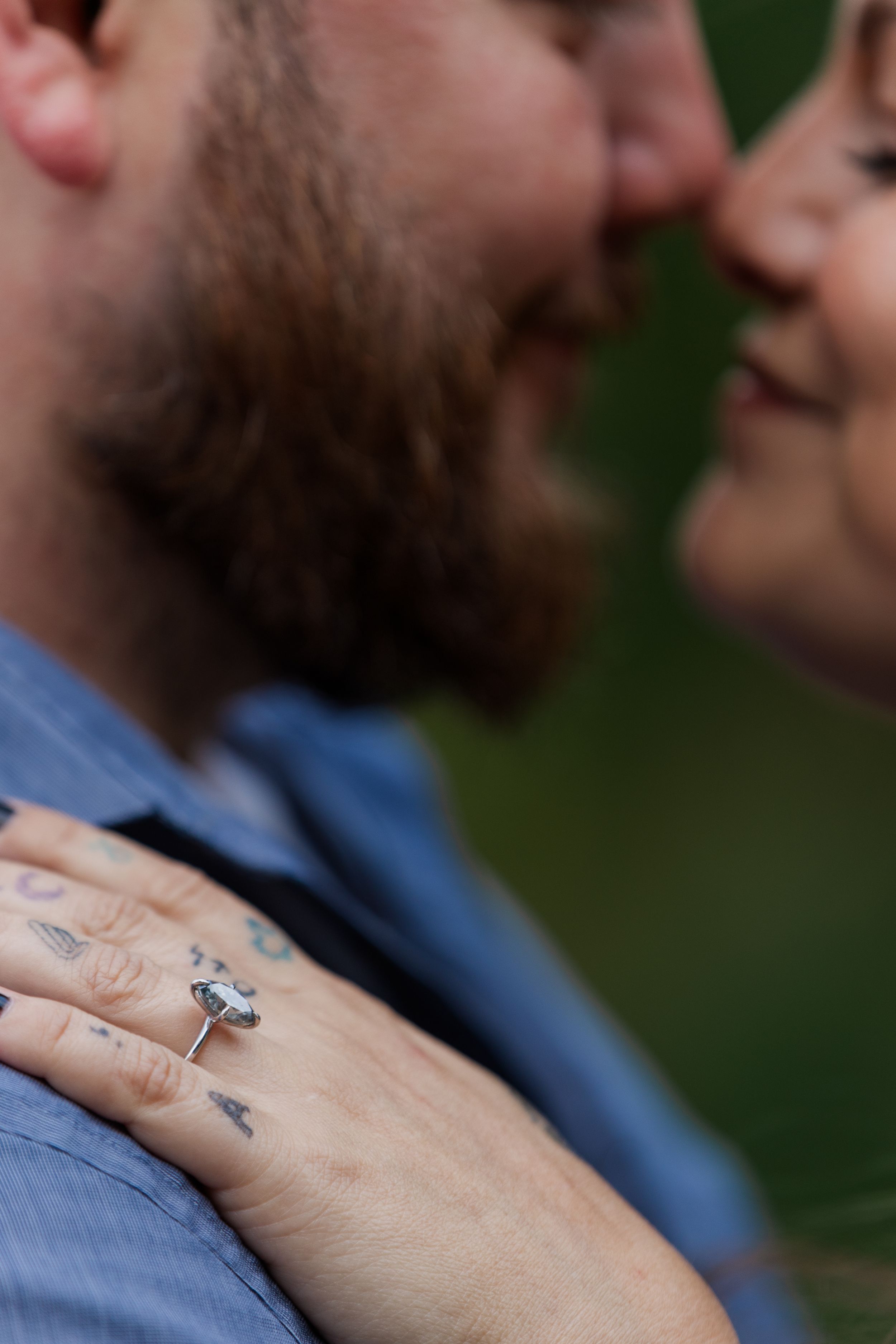 Close-up view of a hand wearing a silver engagement ring against a blurred blue and green background.