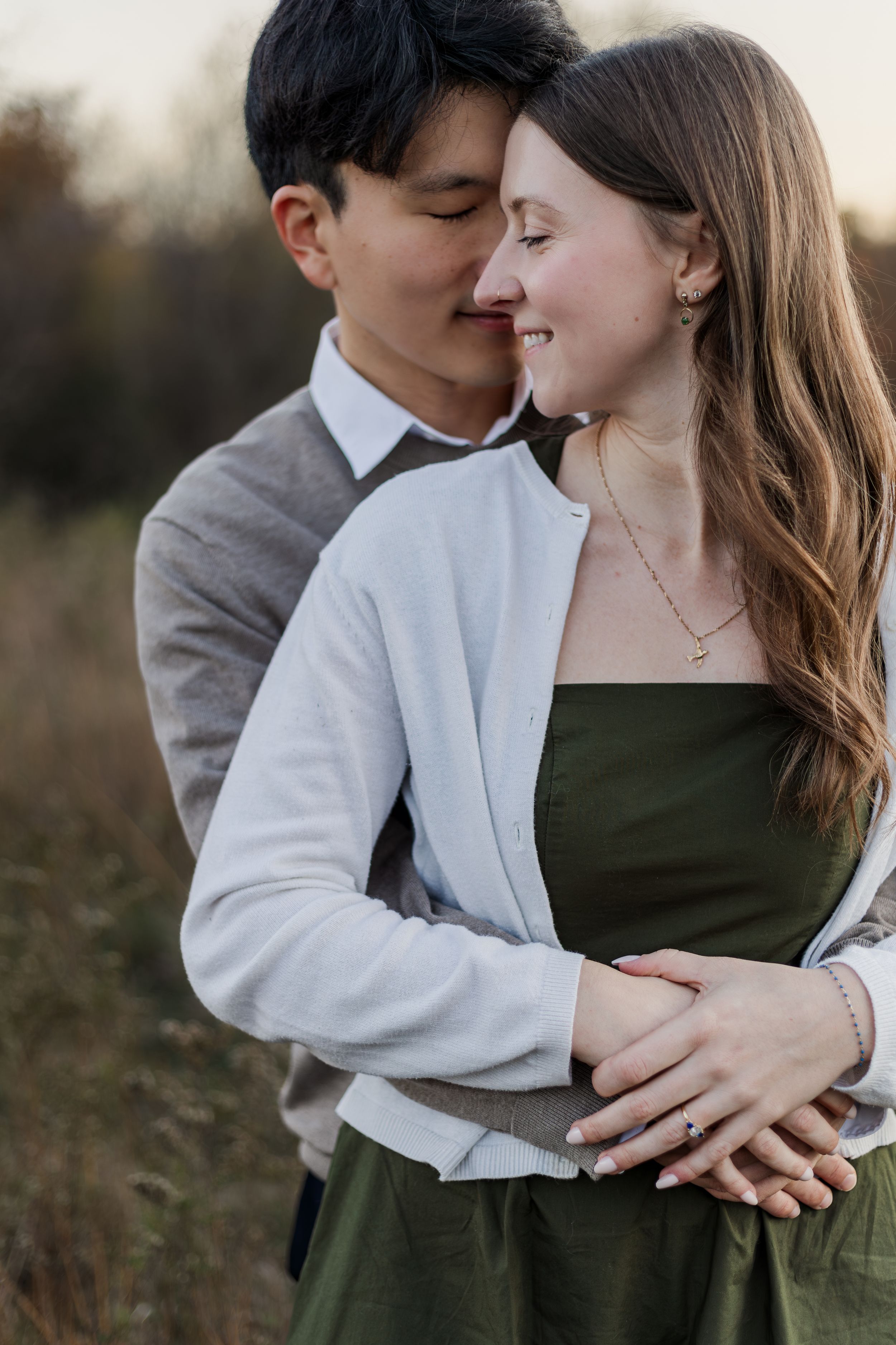 A romantic couple embraces outdoors at sunset, with her wearing a green dress and white cardigan.