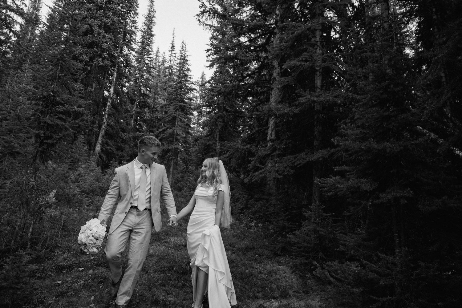 A couple walks hand in hand through a forest of tall evergreen trees on their wedding day.