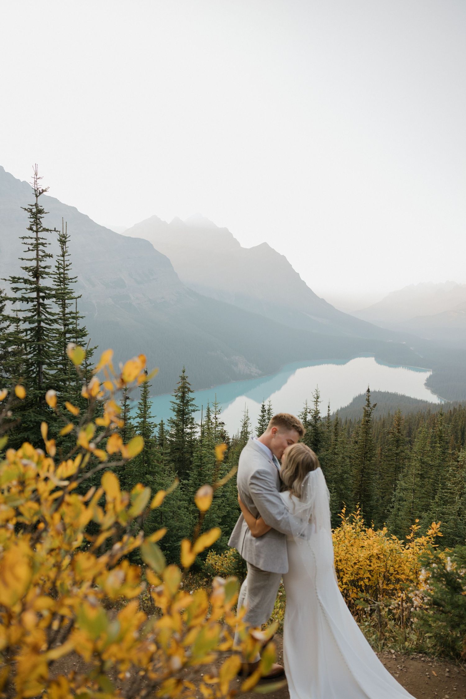 A couple shares a romantic moment at a mountain overlook with a turquoise lake and yellow autumn foliage.