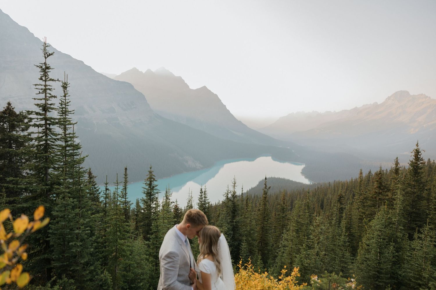 A couple stands on a mountain trail overlooking a scenic alpine lake surrounded by evergreen forest and mountain peaks.