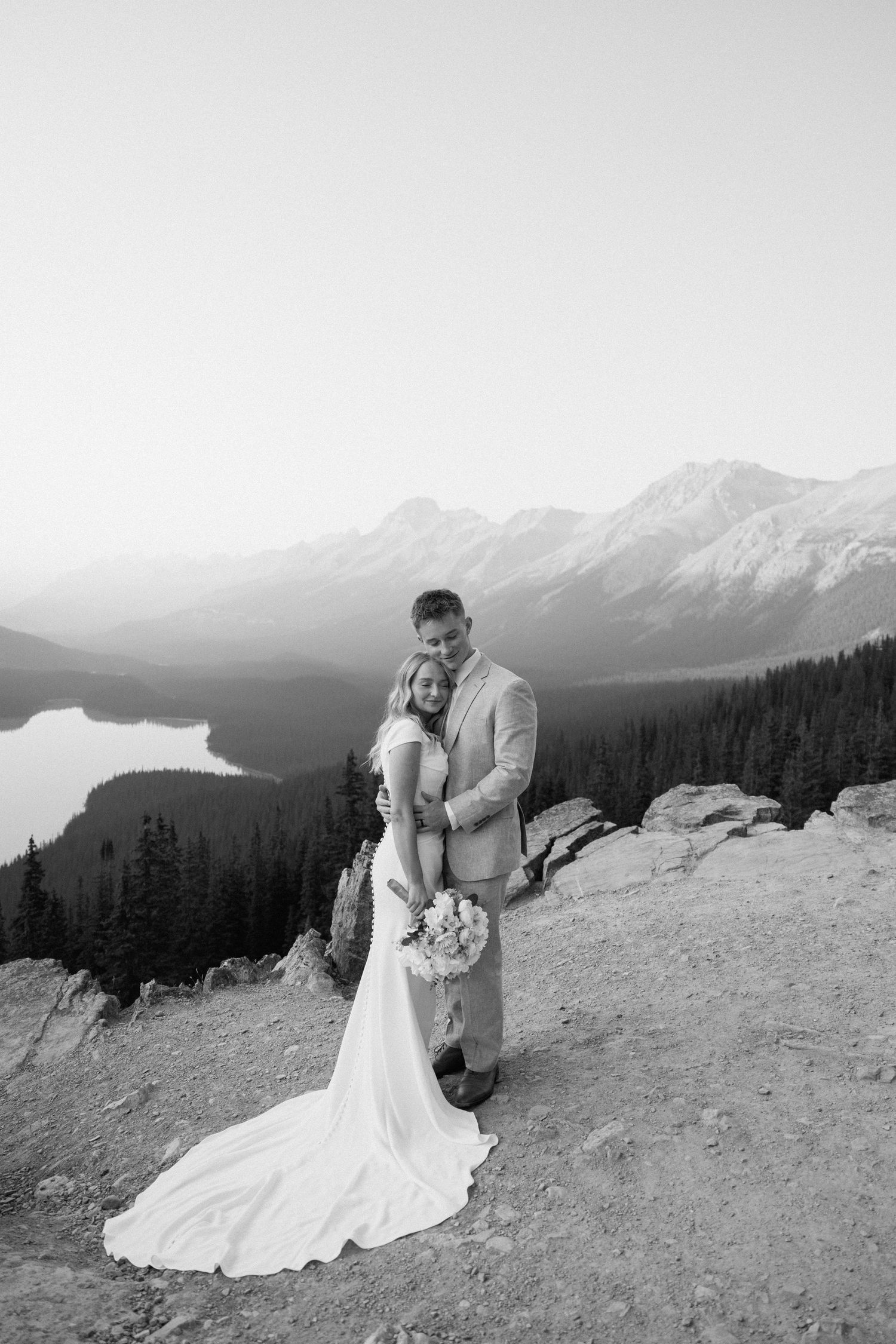A romantic wedding photo captured on a mountain overlook with a scenic lake and peaks in the background.
