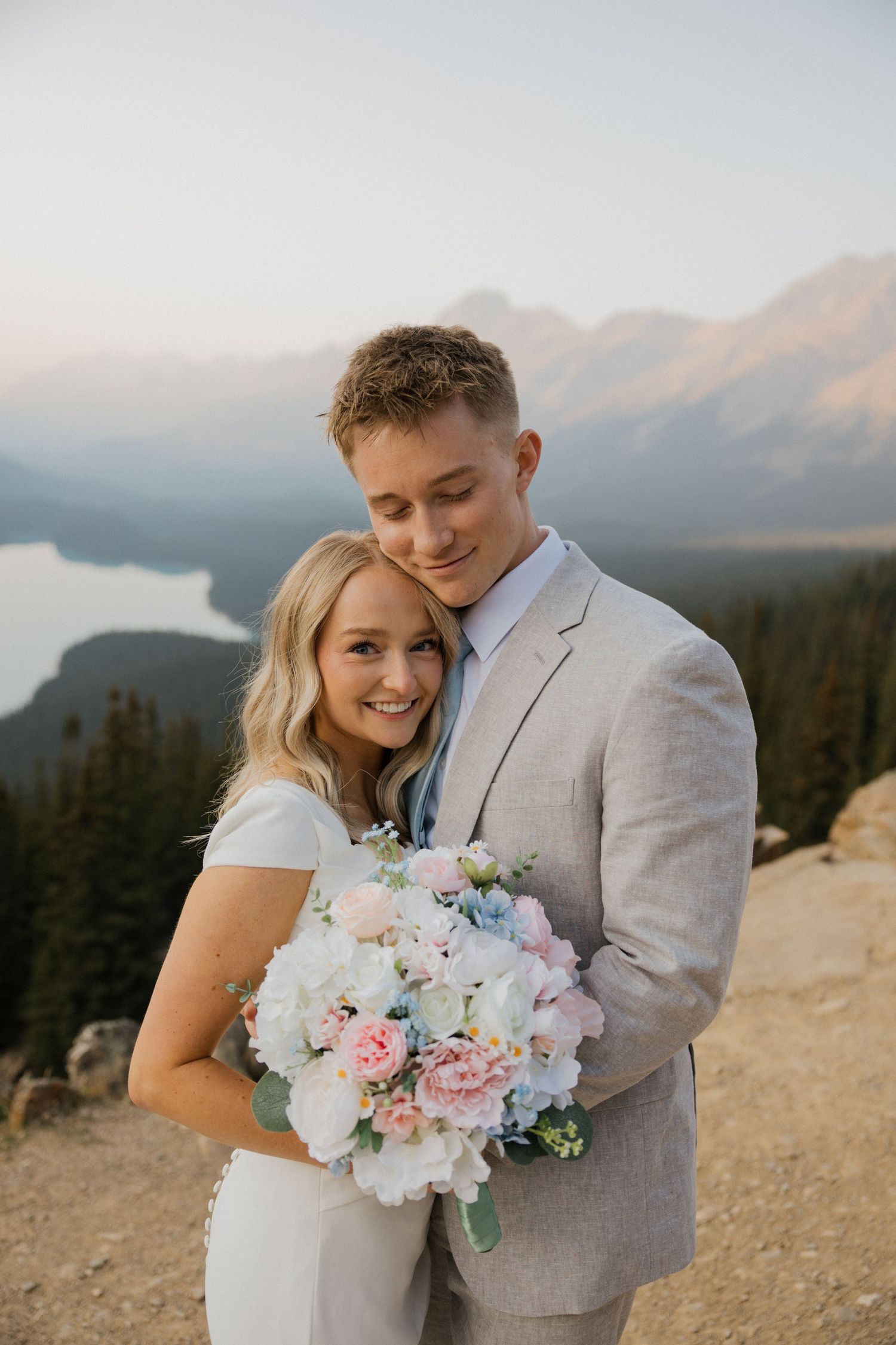 A wedding couple poses on a mountain overlook with a pastel pink and white floral bouquet at sunset.