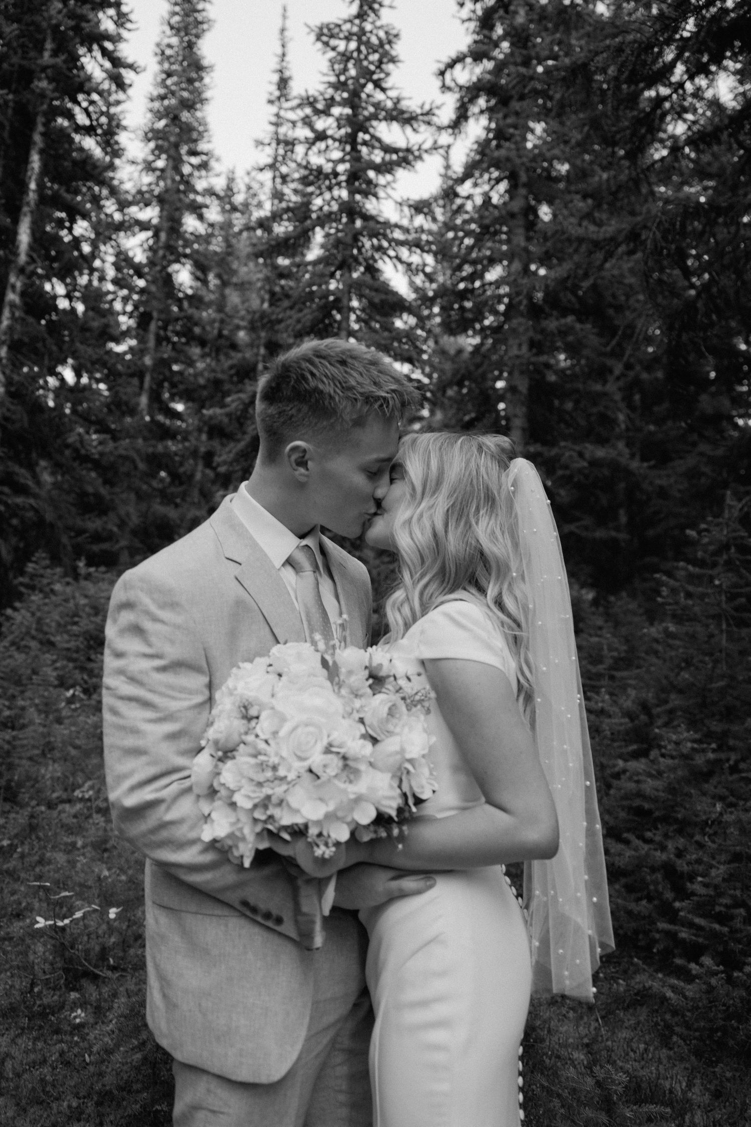 A romantic black and white wedding portrait of a couple kissing in a forest setting with a white floral bouquet.