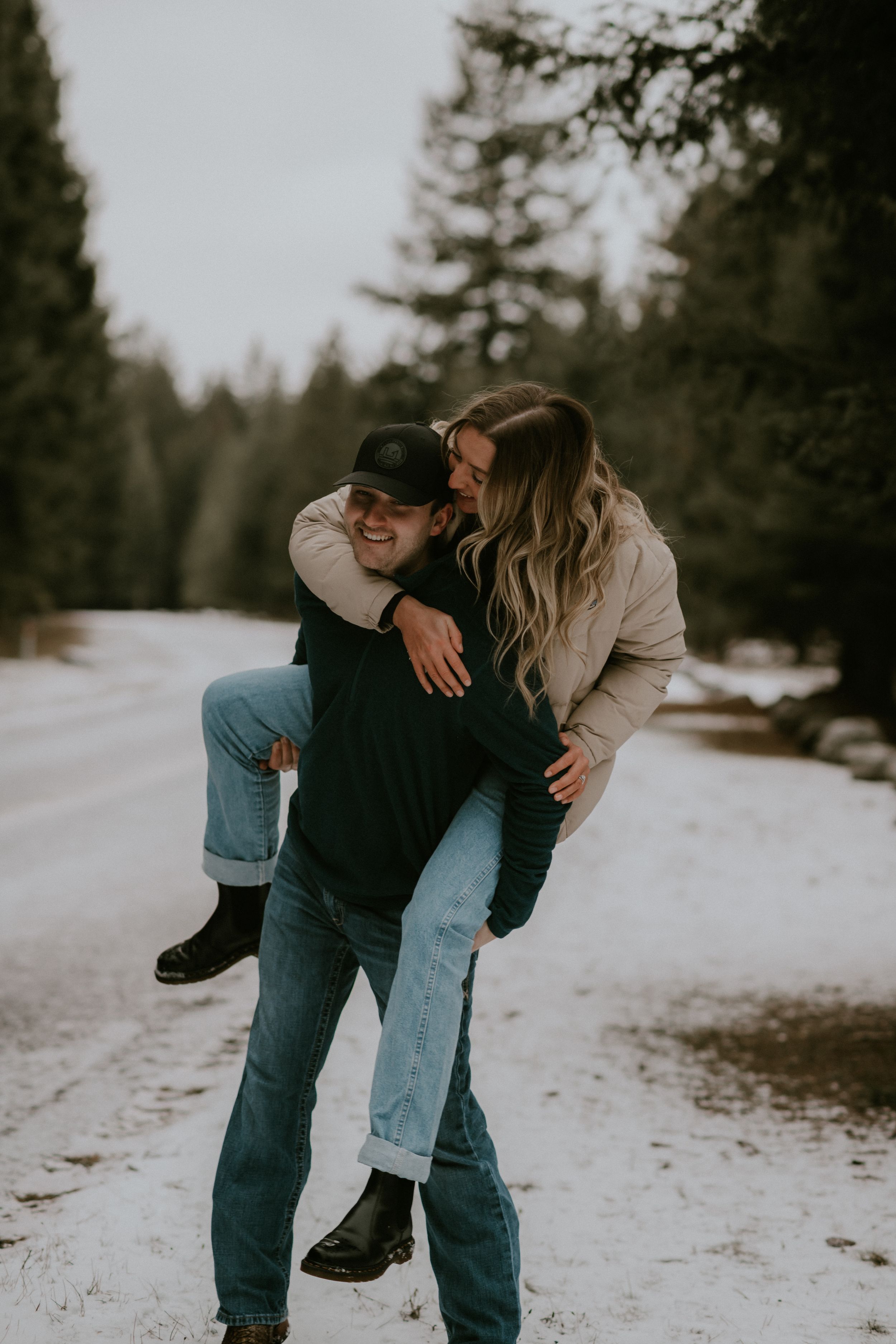 Two people share playful piggyback moments in the snow against a backdrop of evergreen trees.