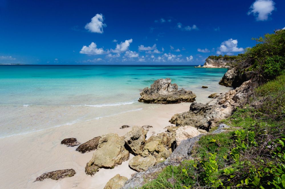 Turquoise Caribbean waters meet white sandy beach with rocky outcrops and lush vegetation under bright blue skies. Landscape.